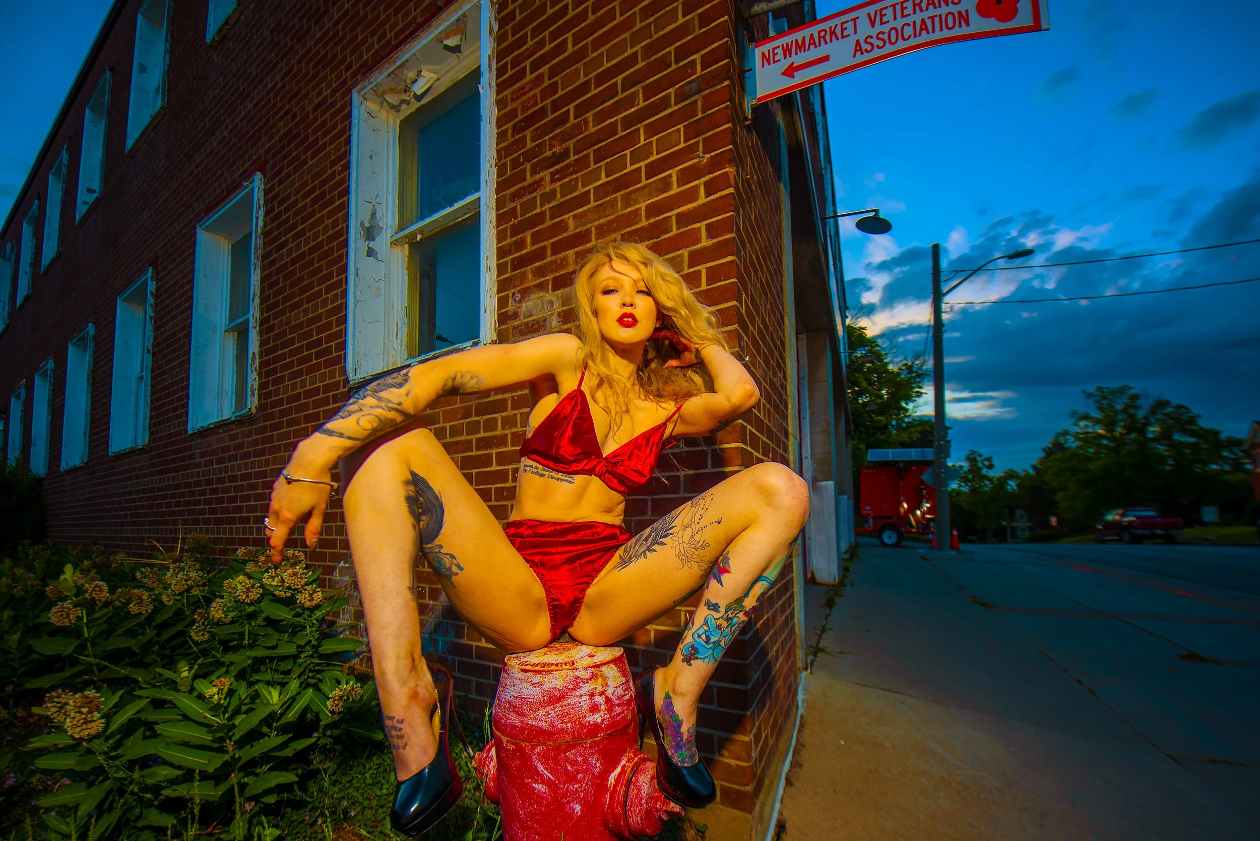 A woman with blonde wavy hair and tattoos posing on a red fire hydrant outside a brick building during dusk, wearing a red satin bra and matching shorts.
