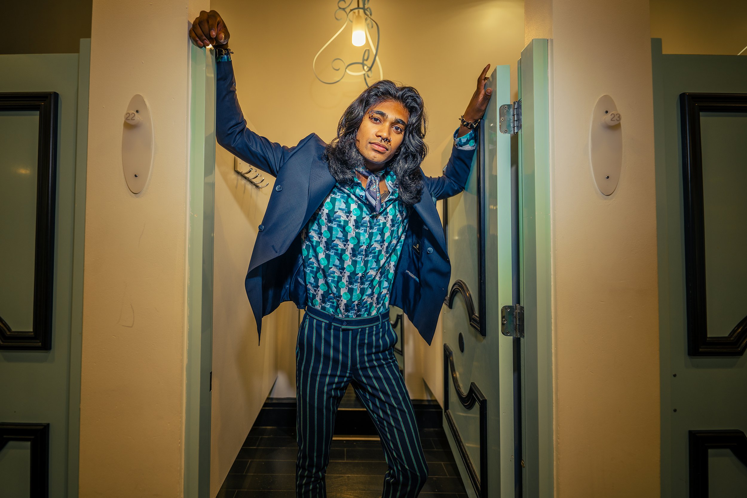 Person with long dark hair and multiple facial piercings posing in a hallway between two doors, wearing a blue patterned shirt, a blue blazer, and striped pants.