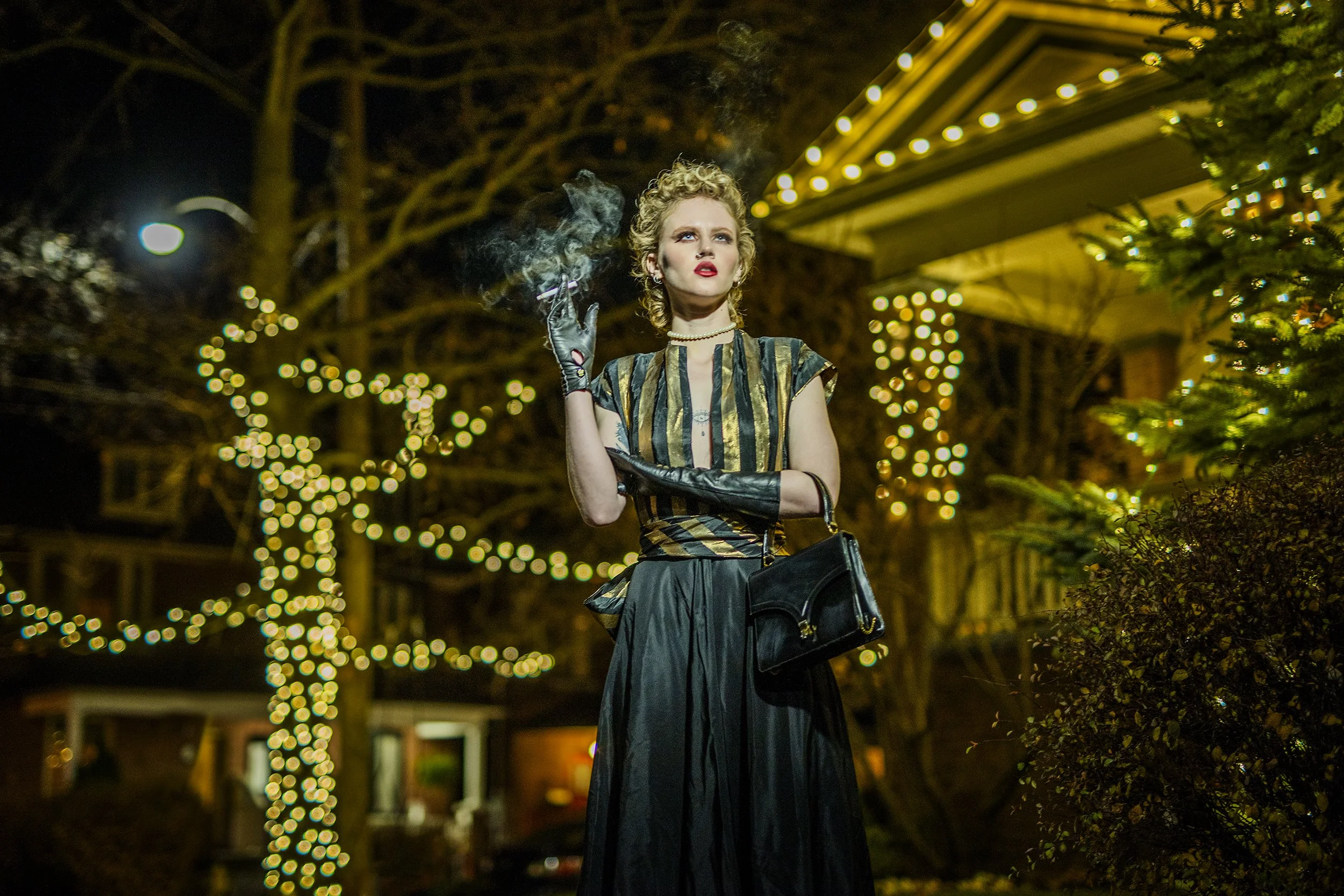 A woman with blonde, curly hair and red lipstick standing outdoors at night near trees decorated with string lights, smoking a cigarette, wearing a striped shirt with metallic accents, and black gloves.