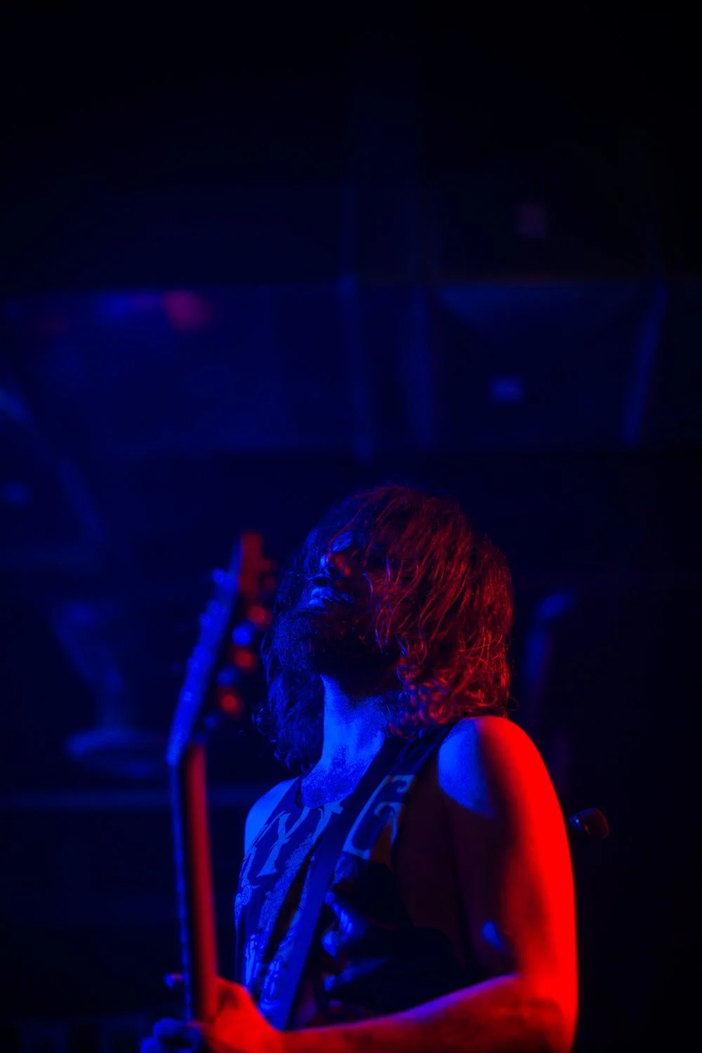 A person with shoulder-length hair playing a guitar under red and blue stage lights in a dark concert setting.