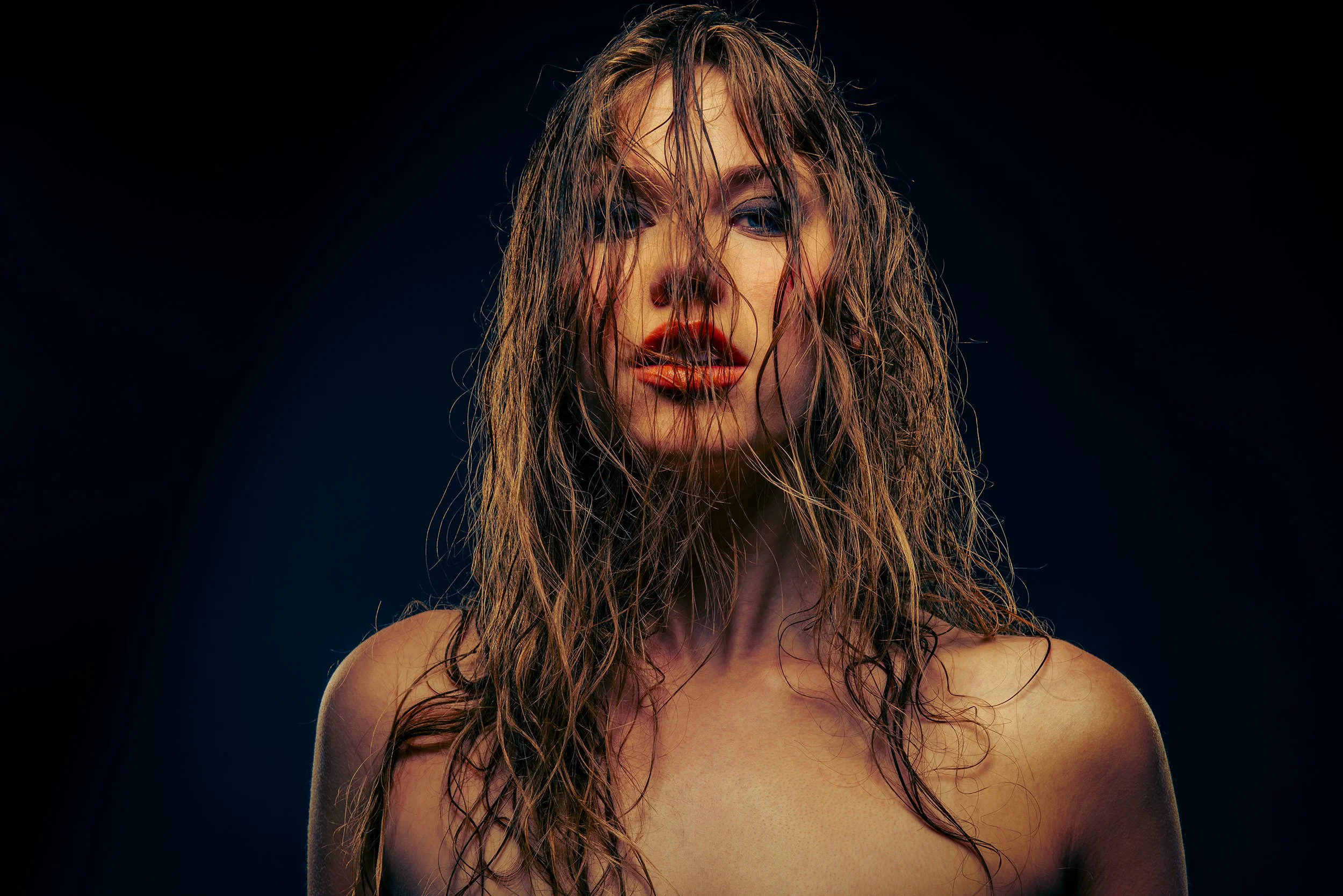 Close-up of a woman with wet, tousled hair and vibrant makeup against a dark background.