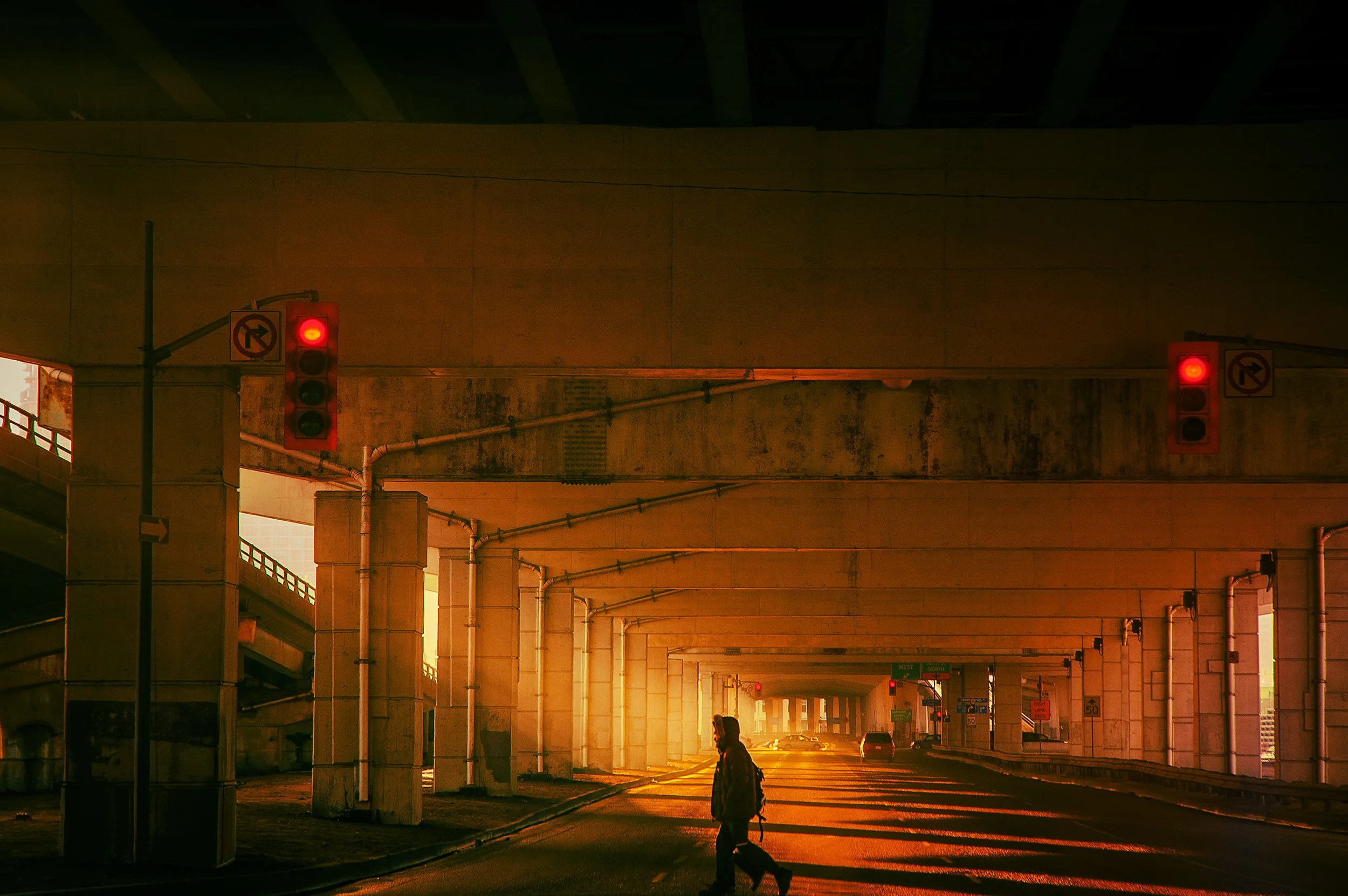 Underpass with red traffic lights glowing, a person walking a dog, and a golden sunset casting shadows, with cars visible in the background.