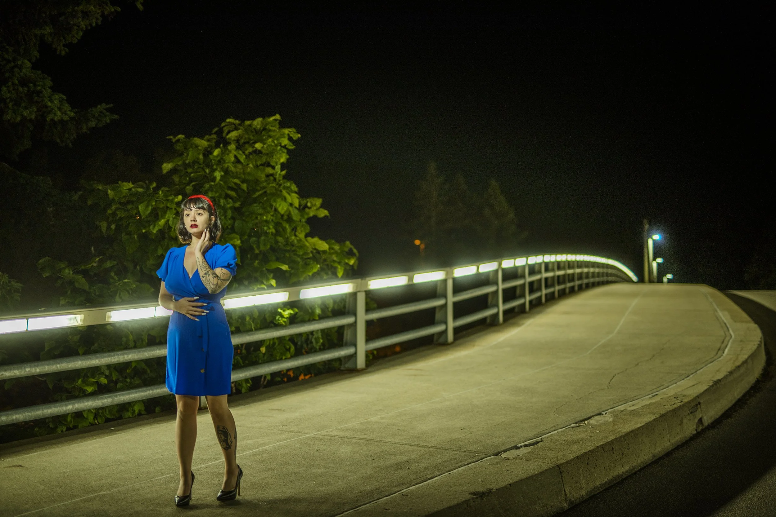 A woman with dark hair, tattoos on her arm and leg, wearing a blue dress and heels, standing on a dimly lit bridge at night, surrounded by greenery and a dark sky.