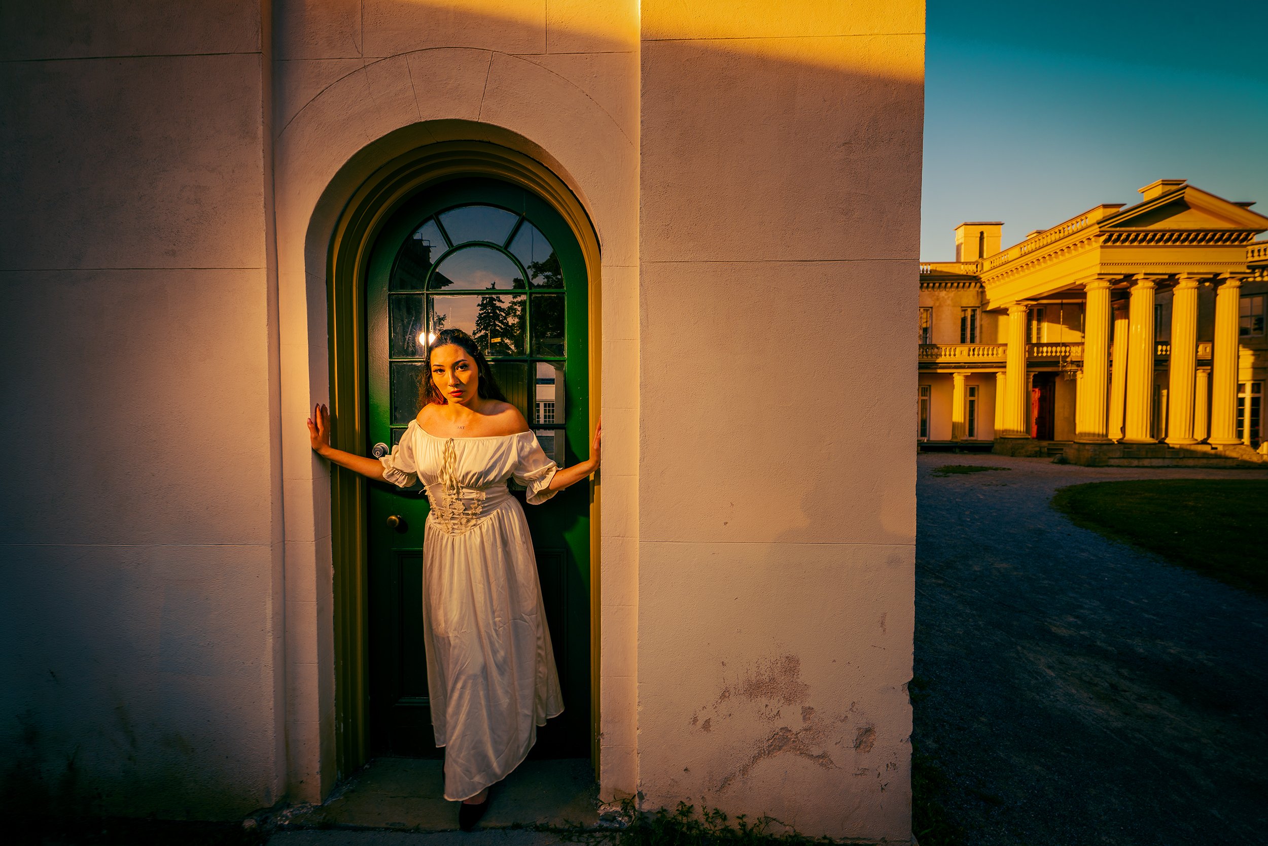 A woman in a white off-the-shoulder dress standing in a doorway of a building during sunset, with yellow light illuminating her and the architectural details in the background.