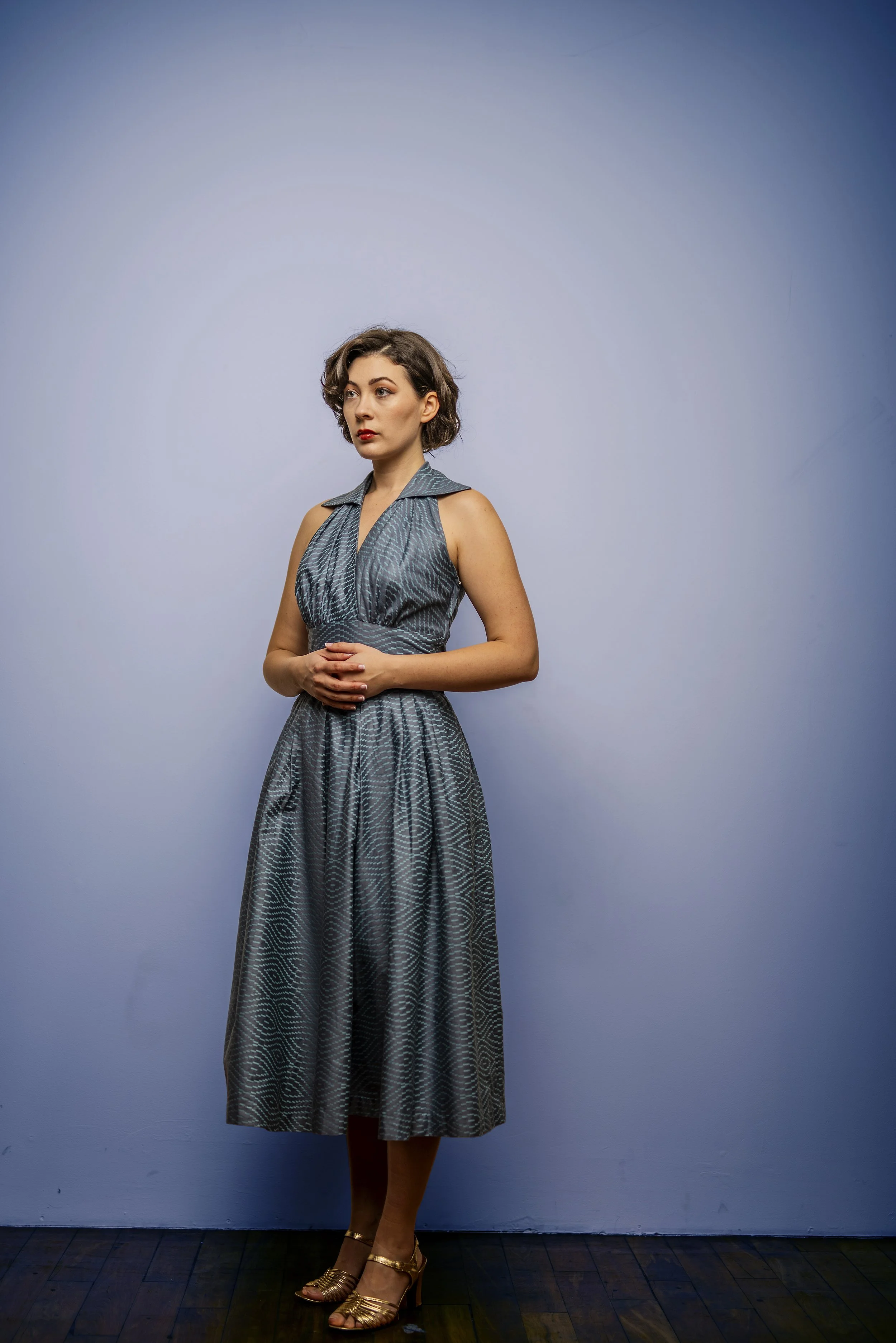 A woman with short, wavy brown hair wearing a sleeveless gray dress with a subtle pattern, standing against a plain light gray background. She has a serious expression and is wearing gold high-heeled sandals.