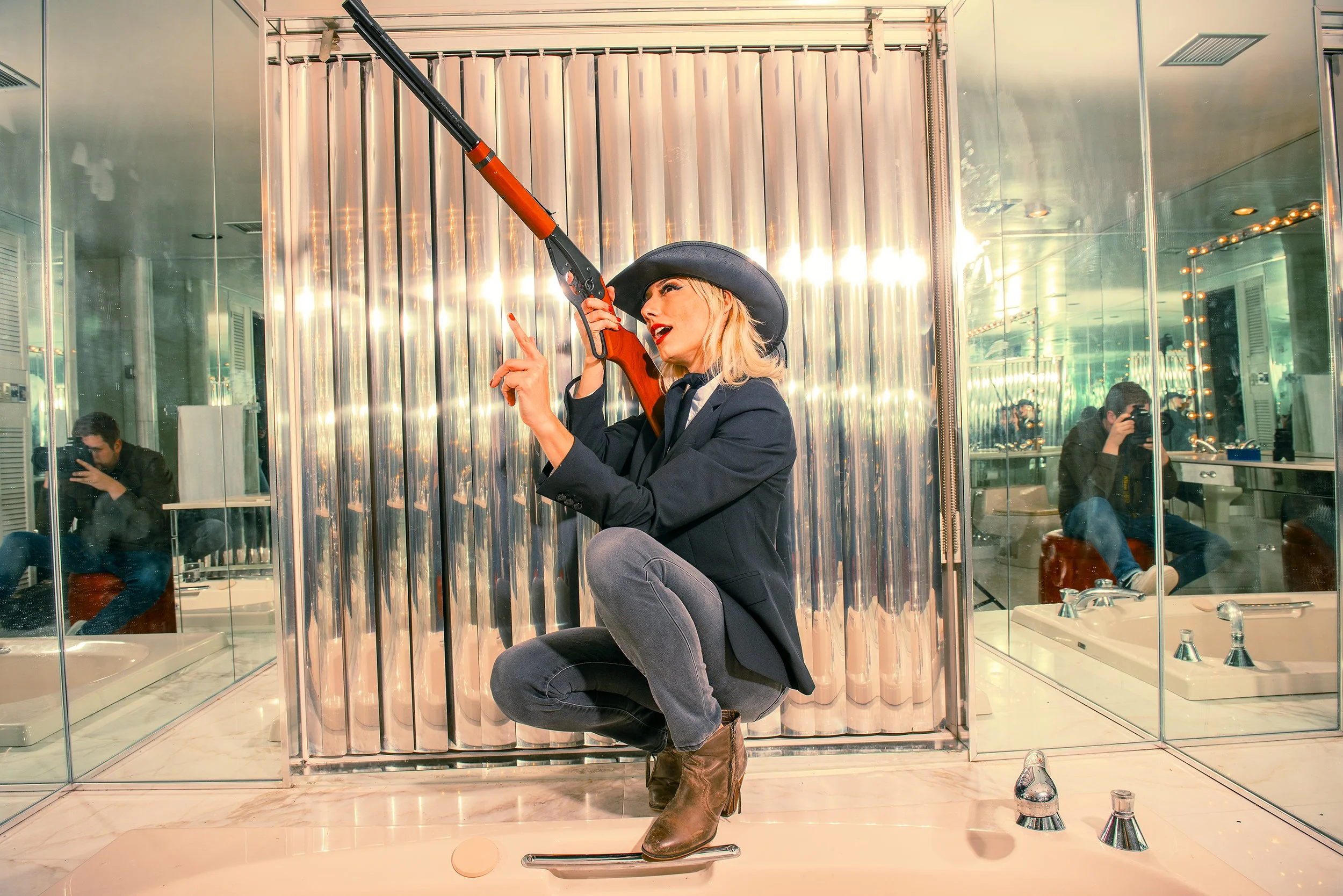 A woman wearing a black cowboy hat and blazer with gray pants and brown boots is squatting inside a mirror bathroom, holding a rifle, with a woman taking a photo in the background. The bathroom has reflective walls and a sink with bottles, and a marb