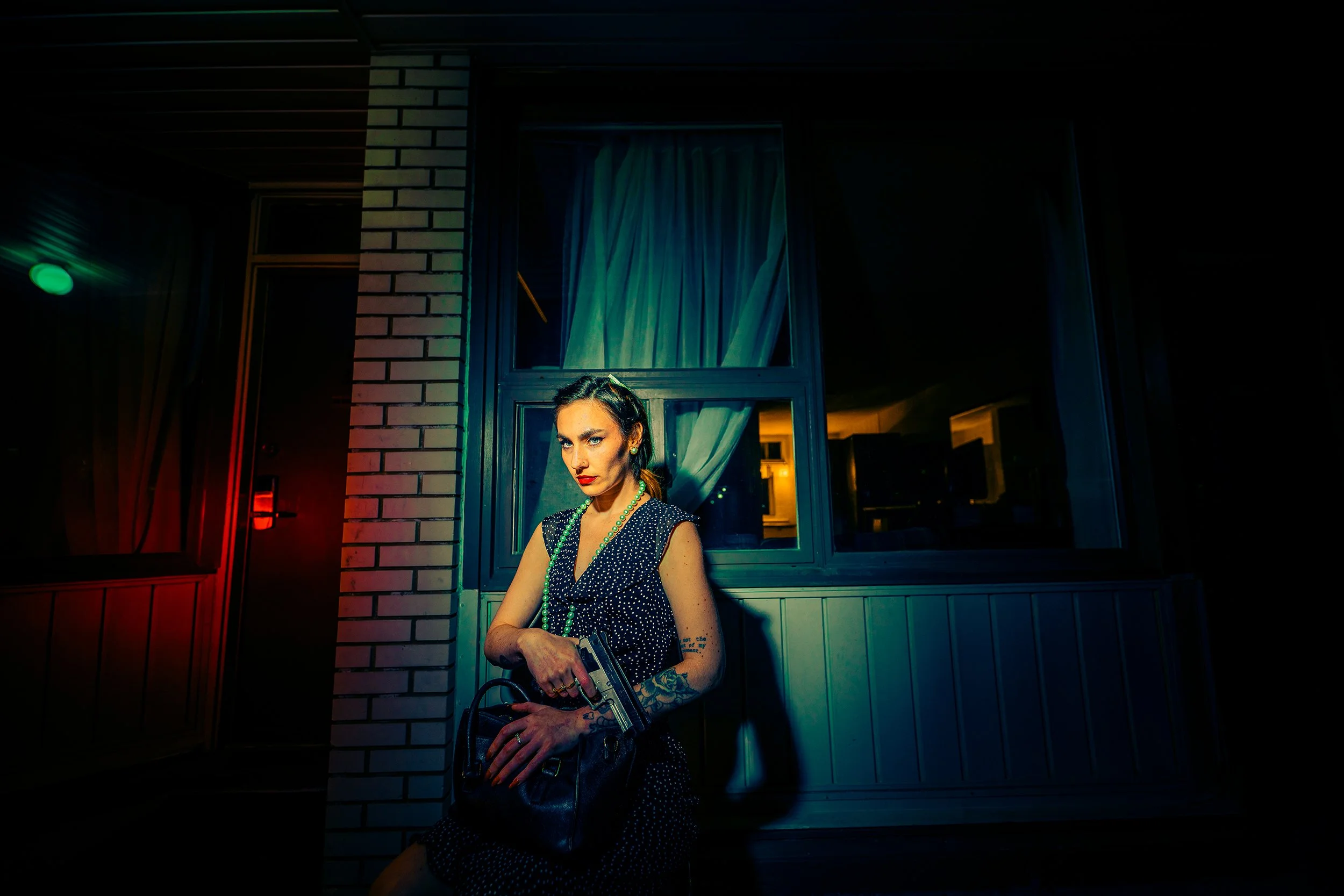A woman with tattoos in a vintage polka-dot dress and green beads, standing outdoors at night, holding a handbag, with a brick wall and window behind her, illuminated by colorful neon lights.