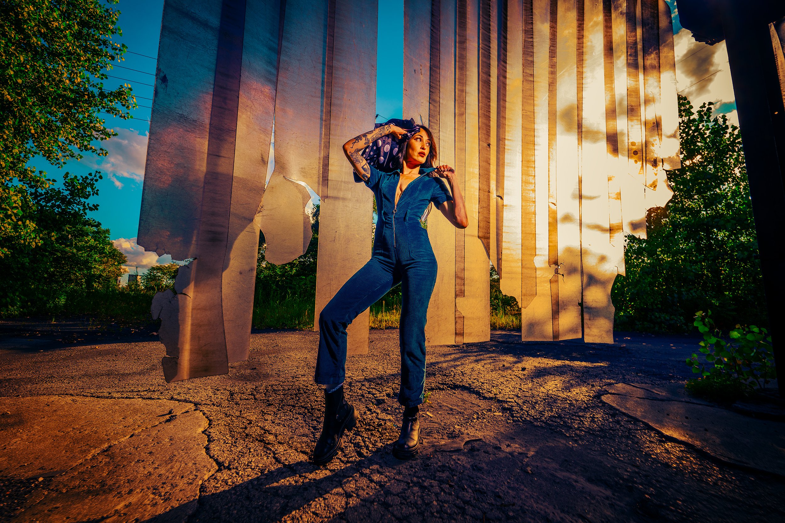 A woman with tattoos on her arms and dark hair, dressed in a blue jumpsuit and black boots, standing outdoors at sunset near an art installation of large, hanging, reflective metal sheets. She is looking into the distance with one hand on her head.