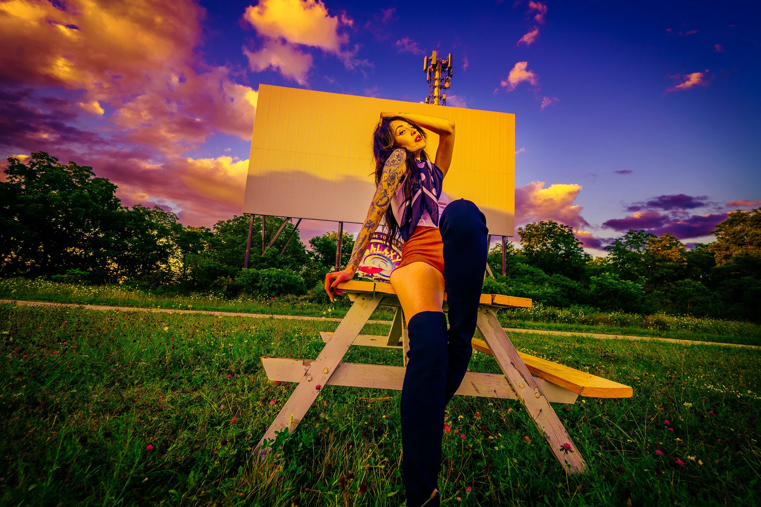 A woman with tattoos posing on a wooden bench outdoors during a colorful sunset with pink, purple, and orange clouds, with trees and a large yellow billboard in the background.