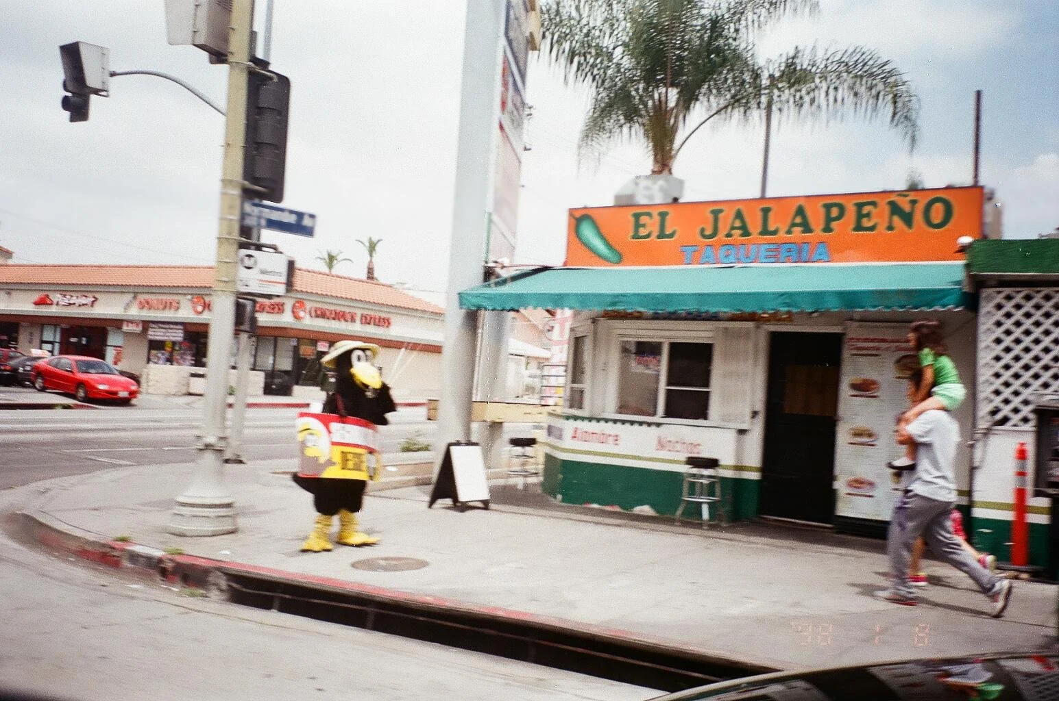 A street corner with a small taco shop called 'El Jalapeño,' with a person carrying a child on their shoulders walking by. A person dressed in a mascot costume resembling a jalapeño with a face, arms, and legs, stands nearby. A palm tree is visible i