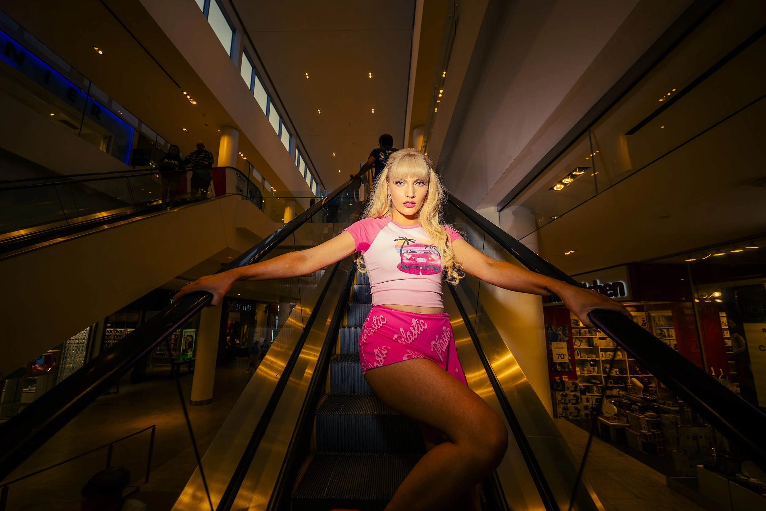 A woman with long blonde hair and makeup, wearing a pink and white graphic T-shirt and pink shorts, poses on an escalator inside a shopping mall.