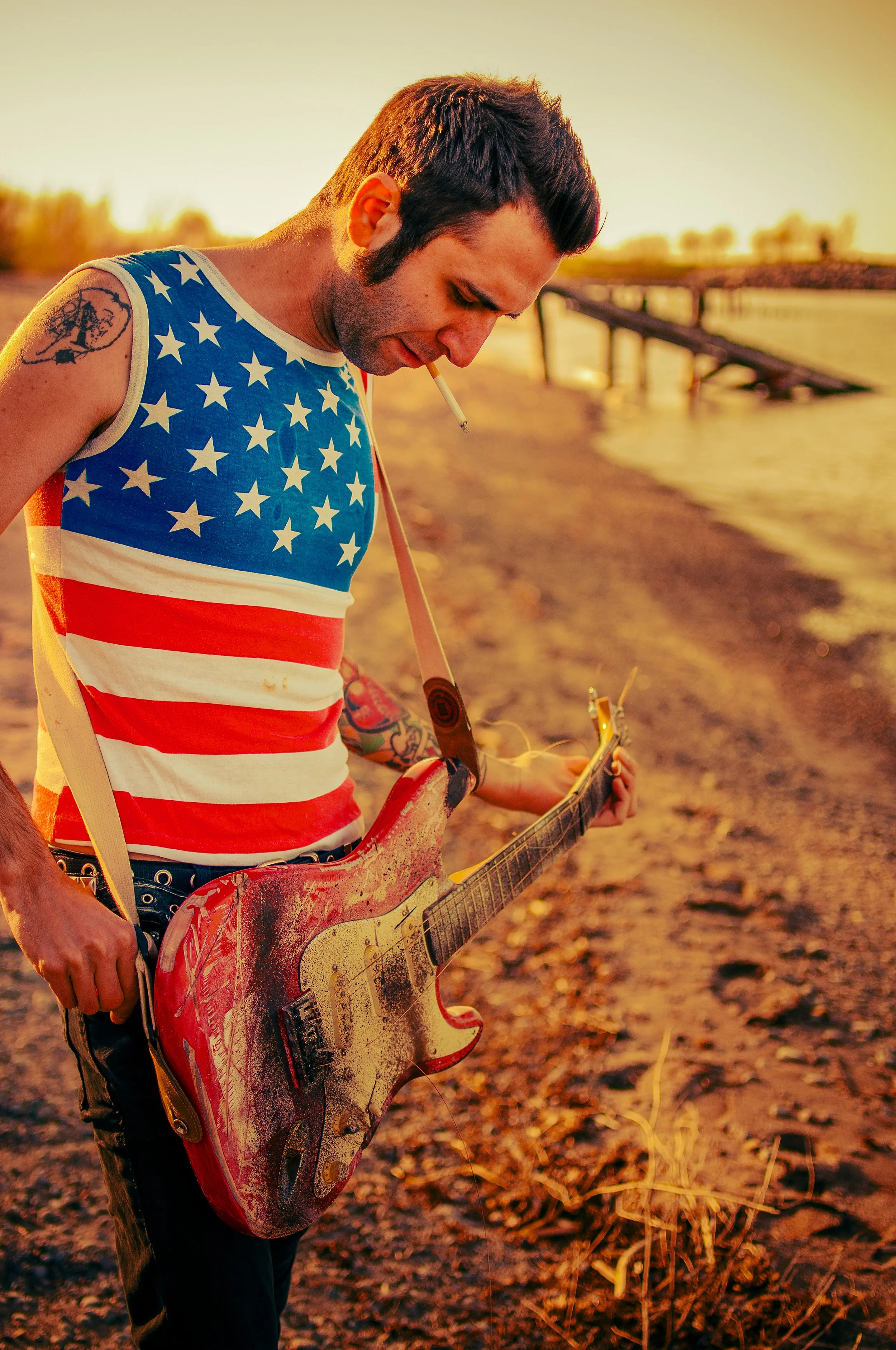 A man with dark hair and tattoos on his arm, wearing a sleeveless tank top with the American flag design, playing a red electric guitar while smoking a cigarette during sunset near a body of water with a bridge in the background.