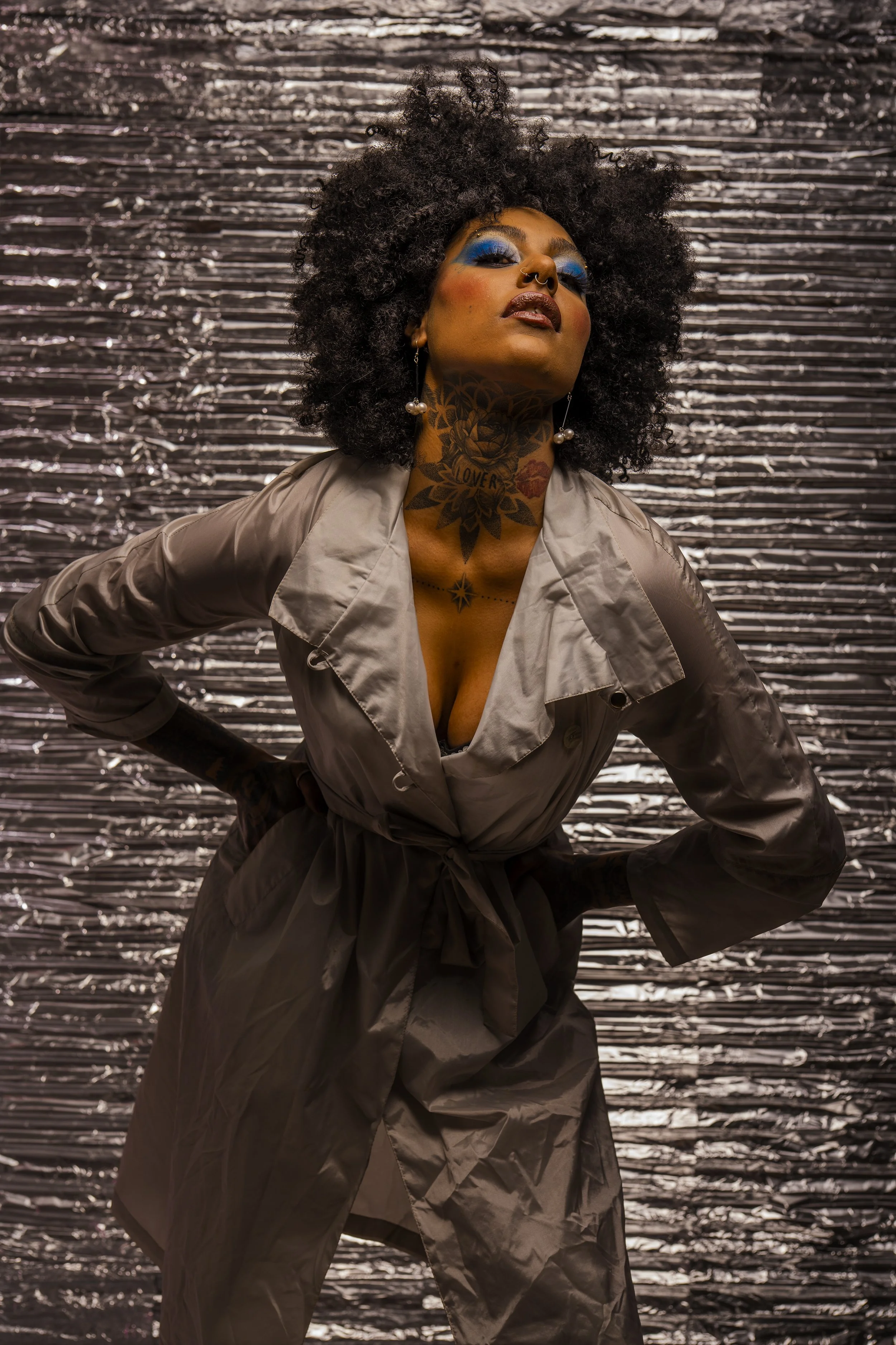 A woman with dark curly hair and tattoos on her neck wearing a beige trench coat, standing with her hand on her hip against a metallic, crinkled background.