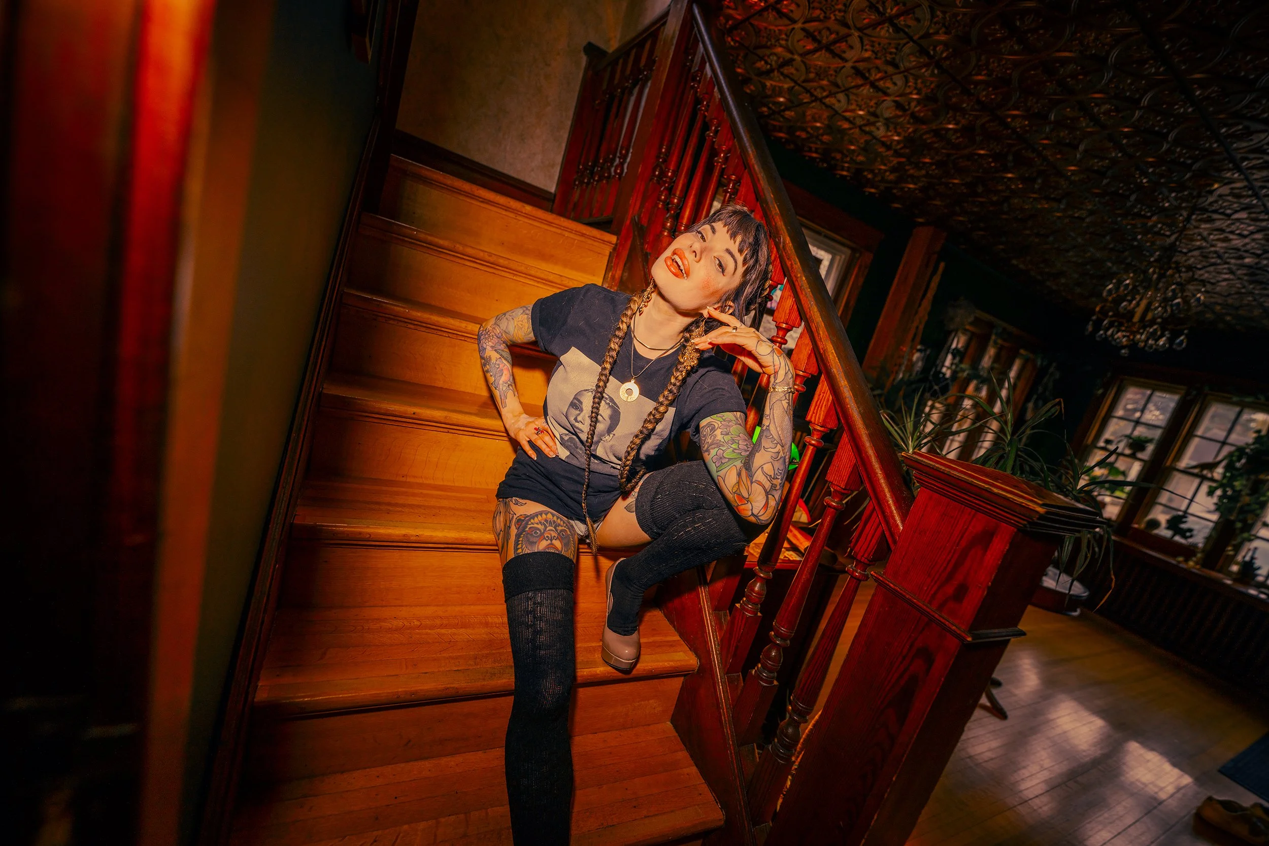 Woman with tattoos sitting on wooden stairs inside a house, tilting her head and smiling.