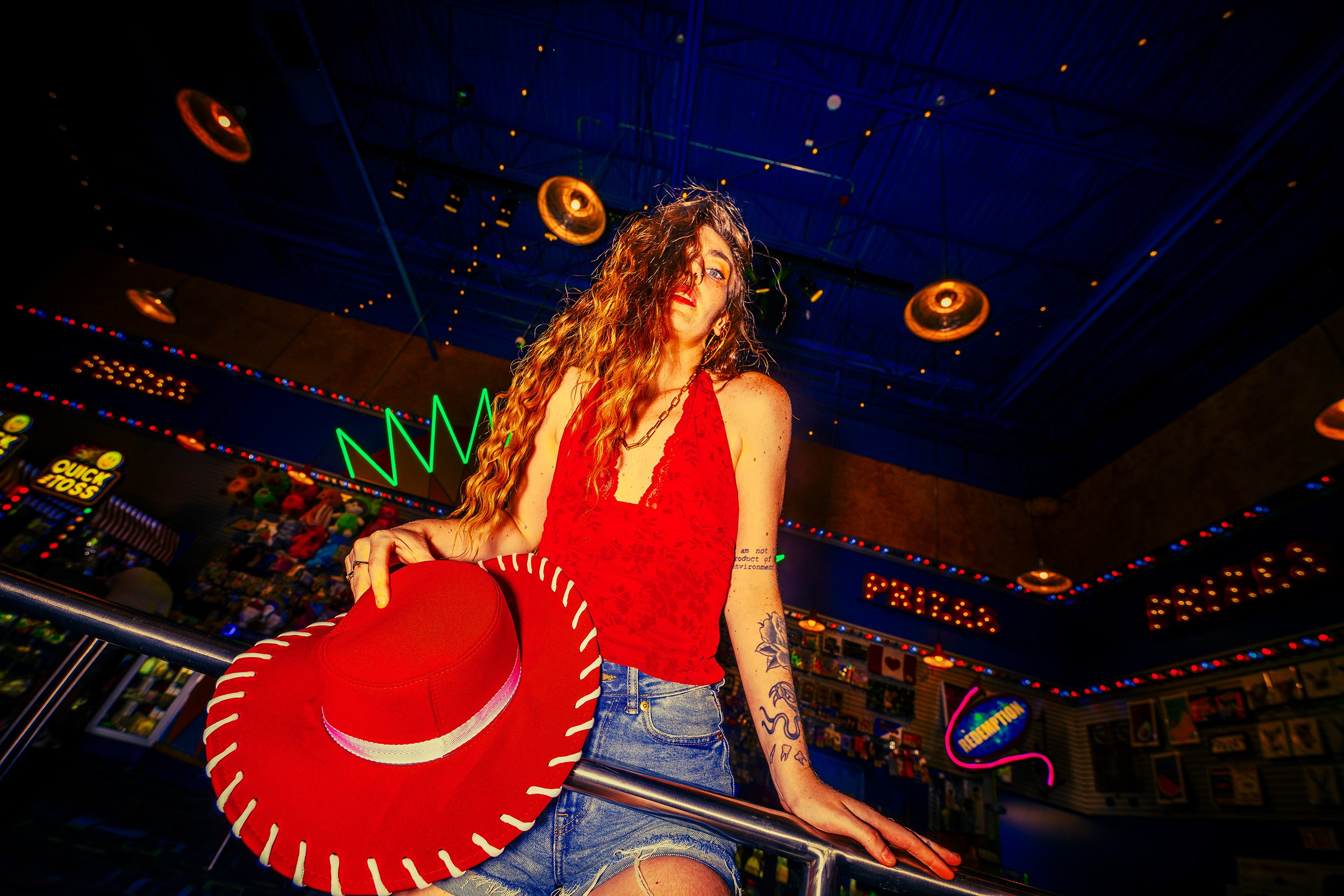 A woman with long curly hair and tattoos on her arm, dressed in a red top and denim shorts, holding a large red hat with white trim inside an arcade or amusement center with colorful lights, game machines, and neon signs.
