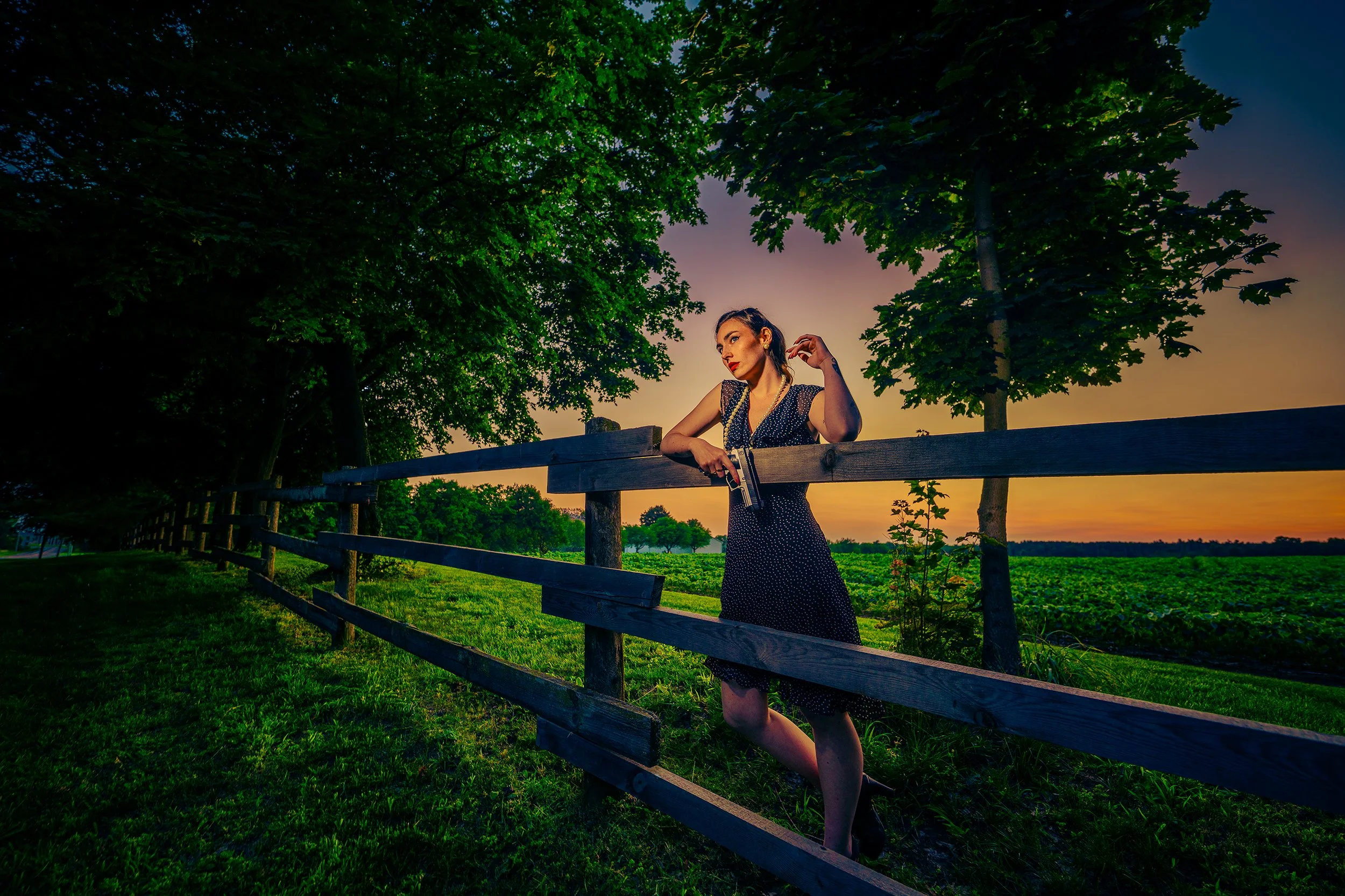 A woman standing behind a wooden fence in a field during sunset, with green trees and a colorful sky in the background.