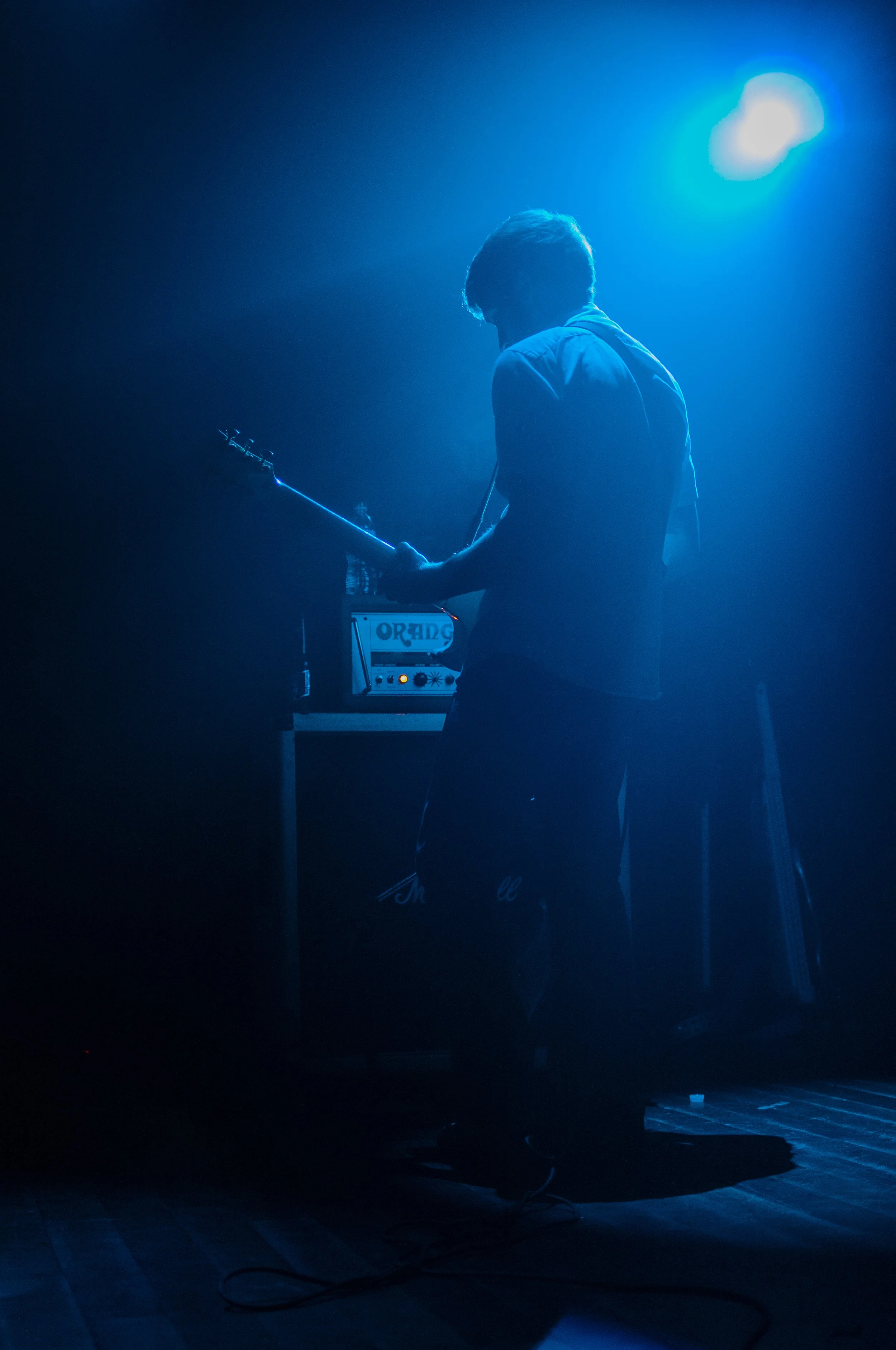 A person with short hair, wearing a hoodie, standing on a dark stage illuminated by blue light, playing an electric guitar, with an amplifier nearby.