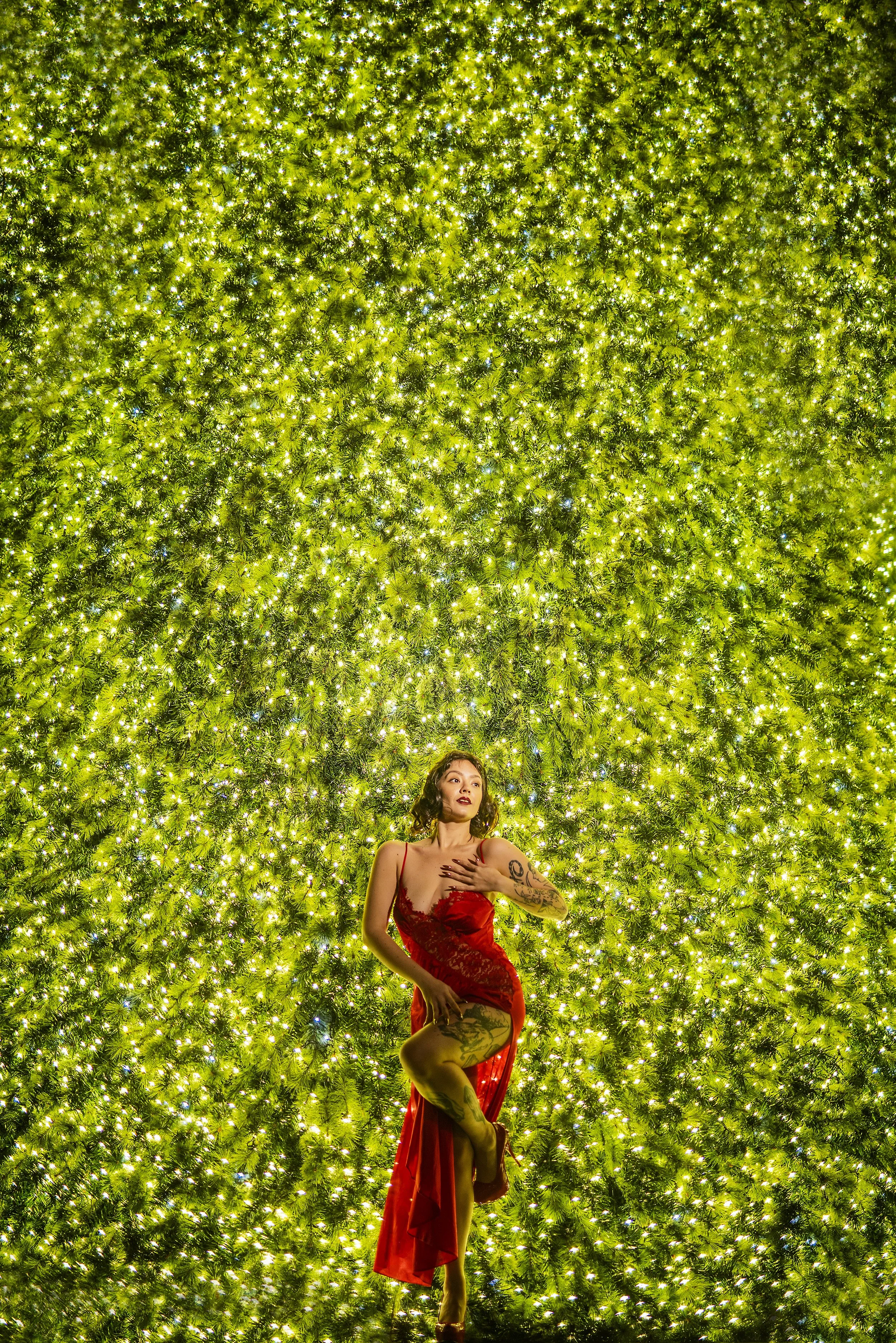 A woman with tattooed arms and legs in a red dress posing in front of a large, illuminated green Christmas tree.