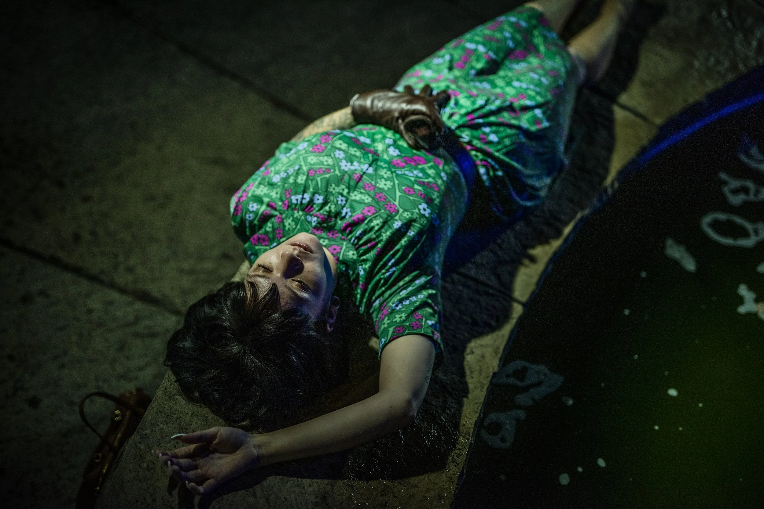 A child lying on a bench at night wearing a colorful floral jacket, eyes closed, with a brown glove covering one hand. The scene is dark with minimal lighting.