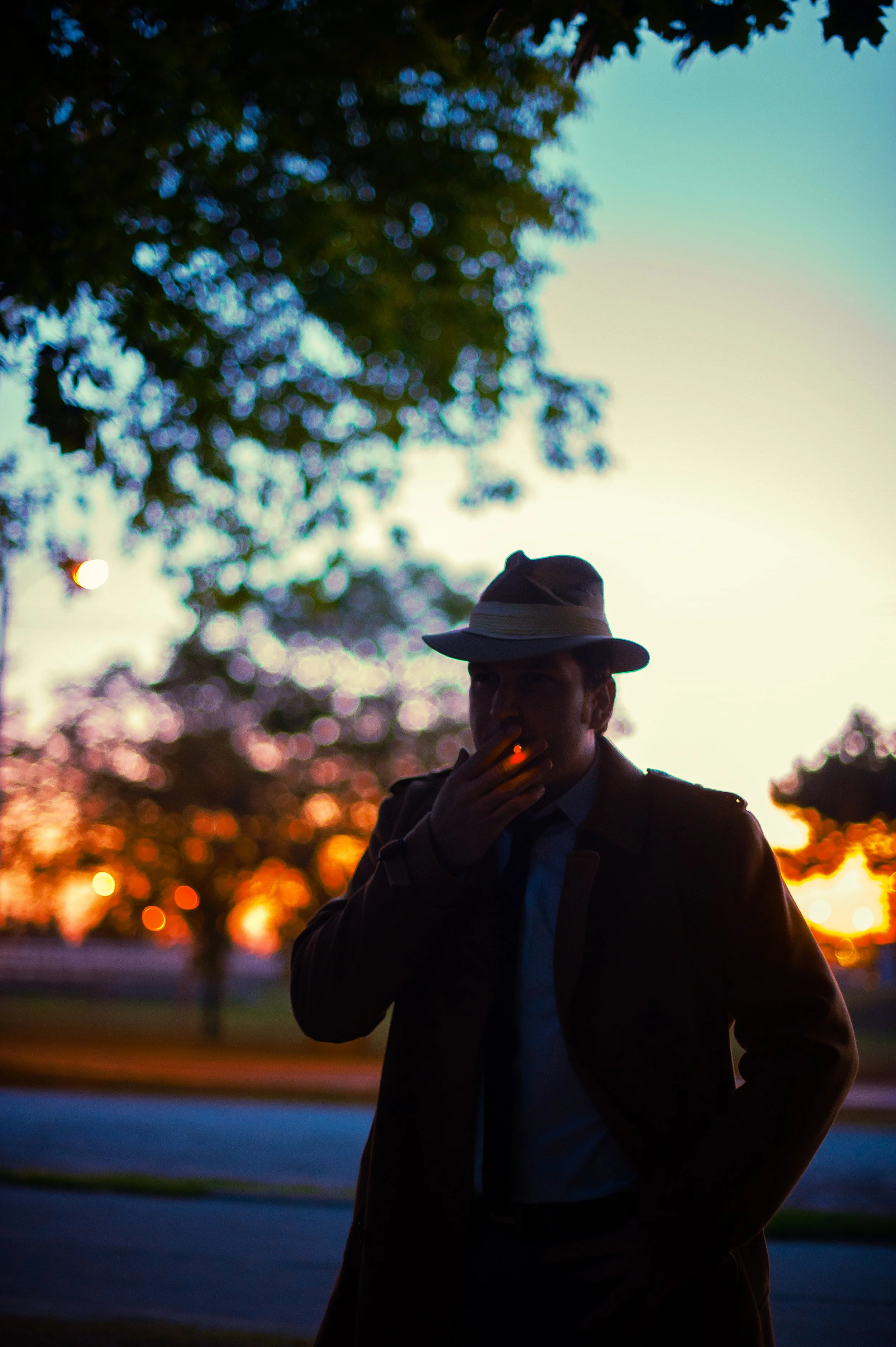 A man wearing a hat and coat, smoking a cigarette, standing outdoors during sunset with a blurred background of trees and a colorful sky.