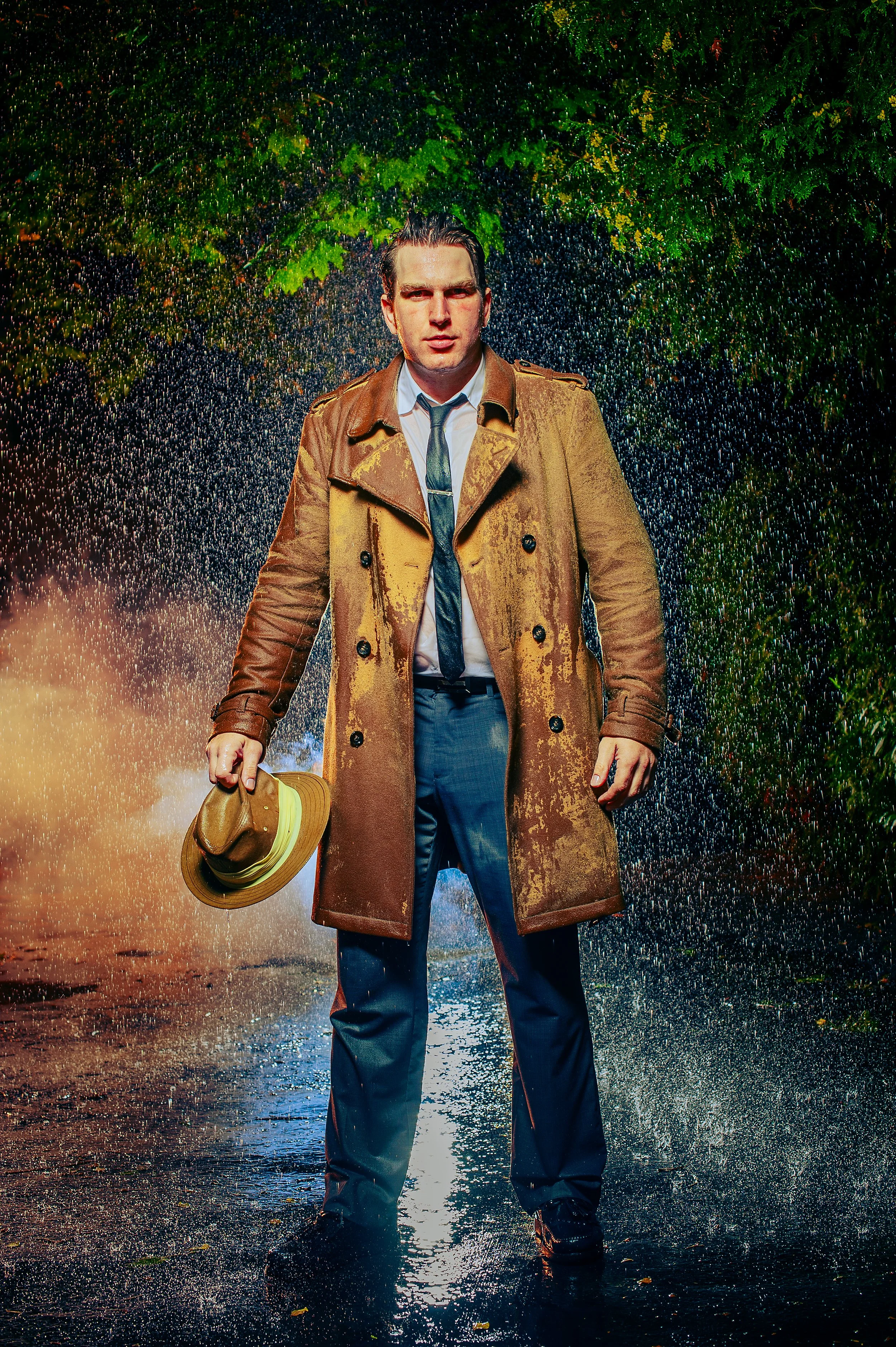 A man in a tan trench coat and blue pants standing in the rain on a wet street, holding a tan fedora. The background features green trees and falling rain.