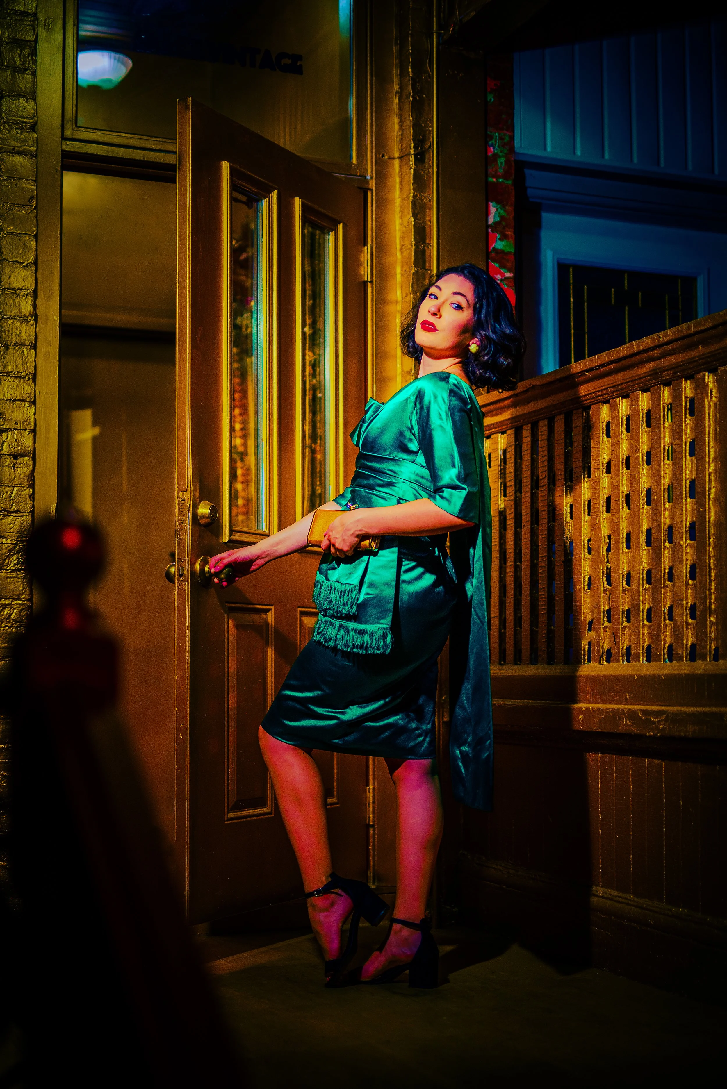 A woman with dark hair, wearing a satin teal dress, standing at a door handle at night, with a wooden fence and brick wall in the background, illuminated by colorful lighting.