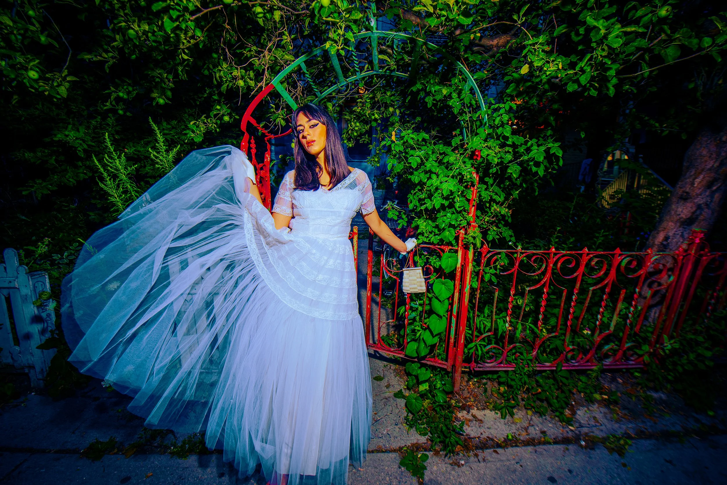 A woman dressed in a white gown holding a white veil standing next to a red decorative fence with lush green foliage behind her.