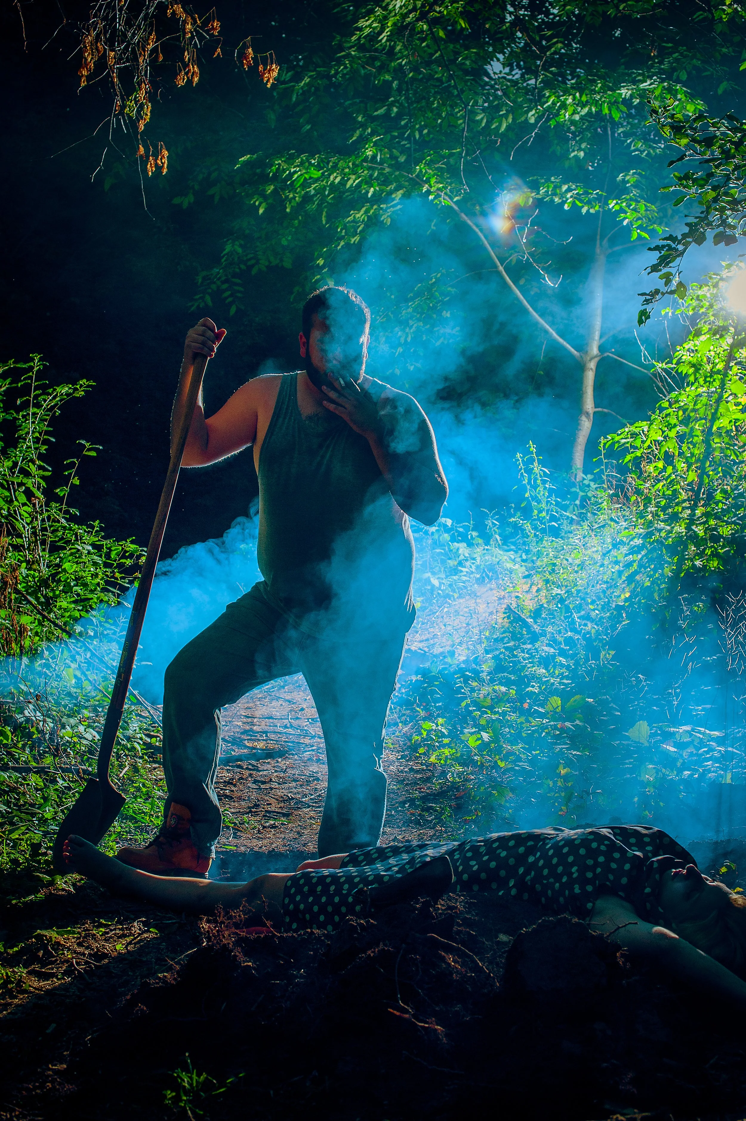 A man kneeling and smoking a cigarette at night in a forested area, with a person lying on the ground nearby, trees and blue smoke surrounding them.