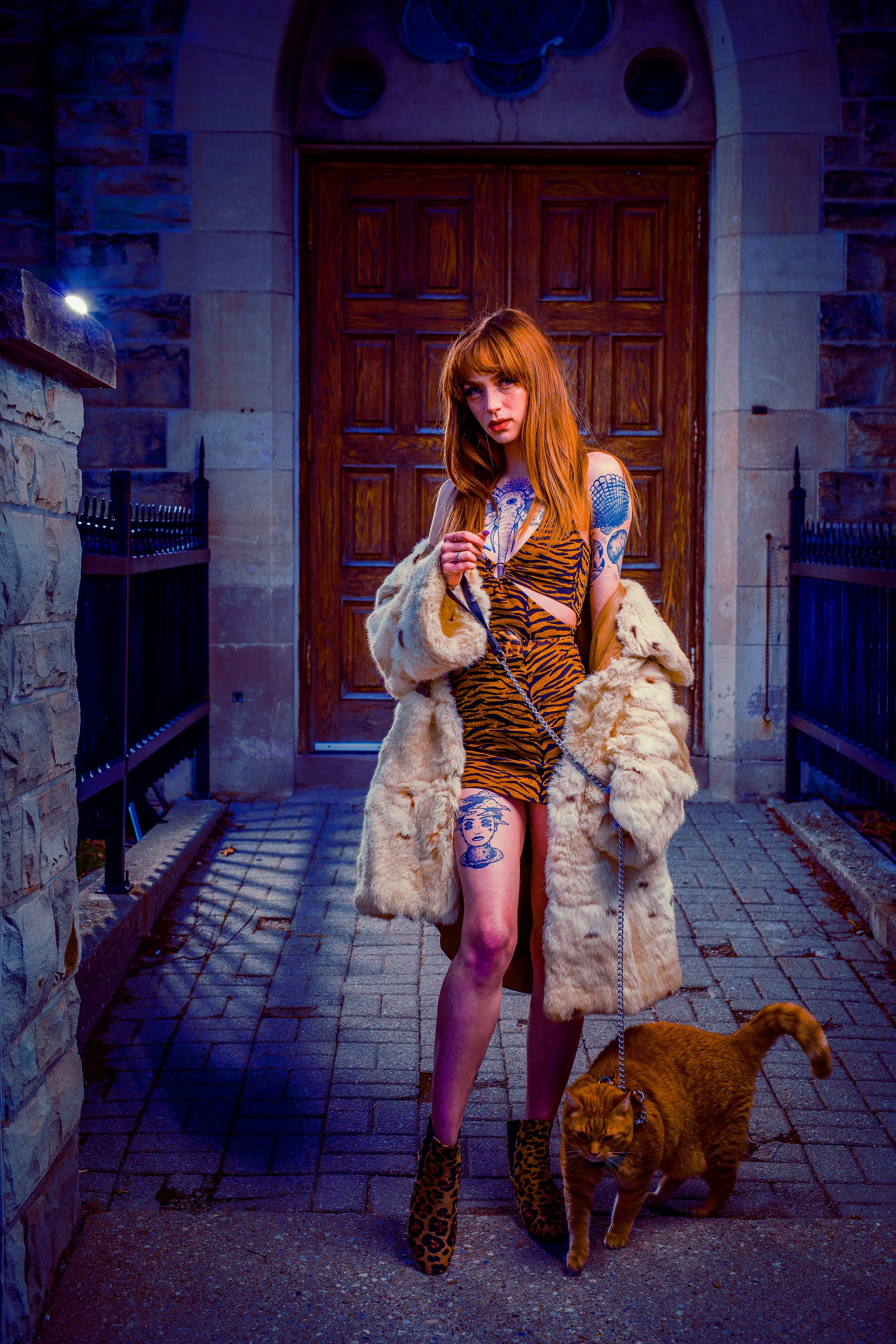 A woman with long red hair and tattoos, wearing a tiger-striped dress and leopard-print boots, walking her brown dog on a city sidewalk at night in front of a wooden door and stone building.