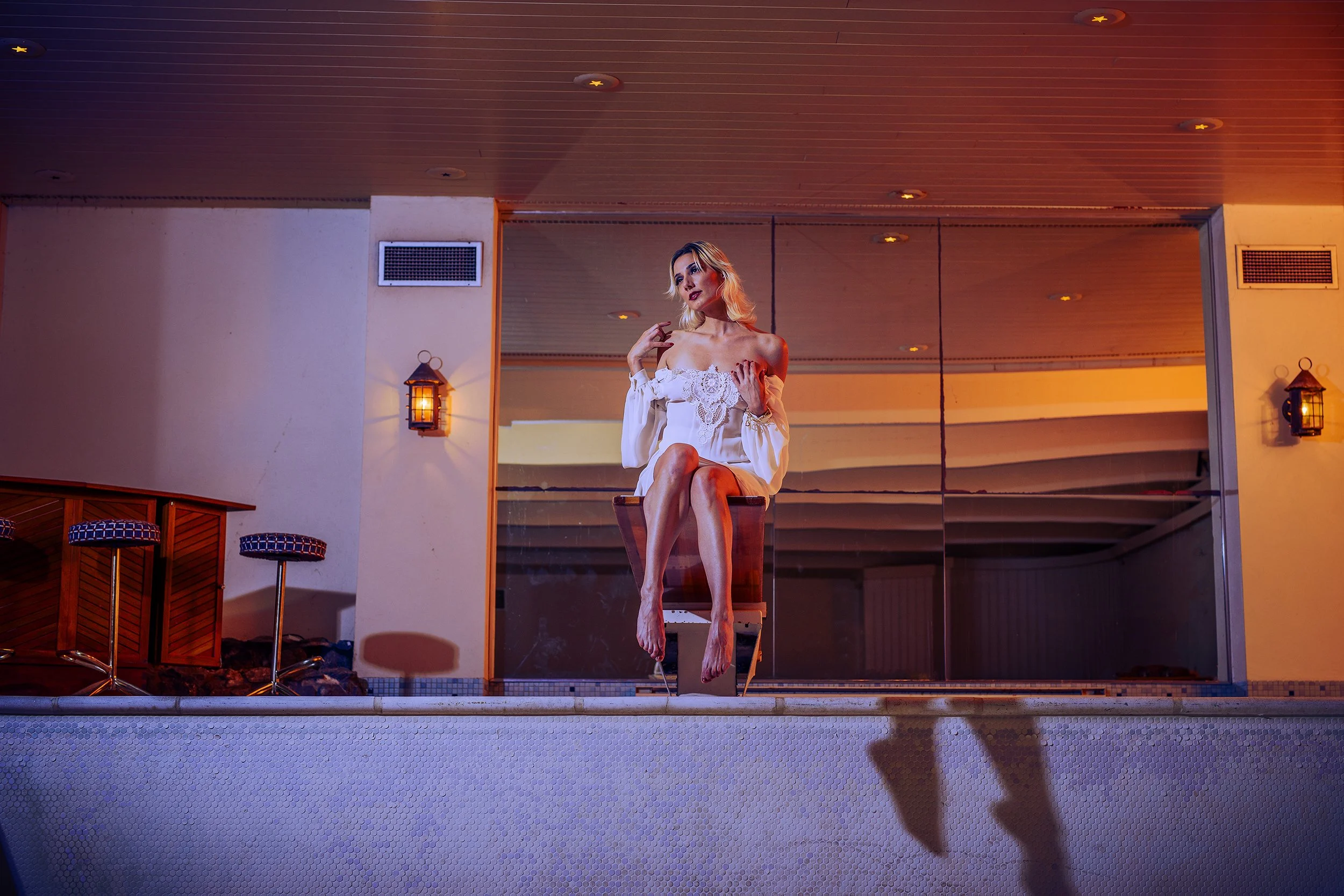 A woman with blonde hair seated on a chair, wearing an off-the-shoulder white dress with lace details, with her legs crossed and holding her upper arm. The setting is indoors with warm lighting, and there are lantern-style wall lights, a mirror behin