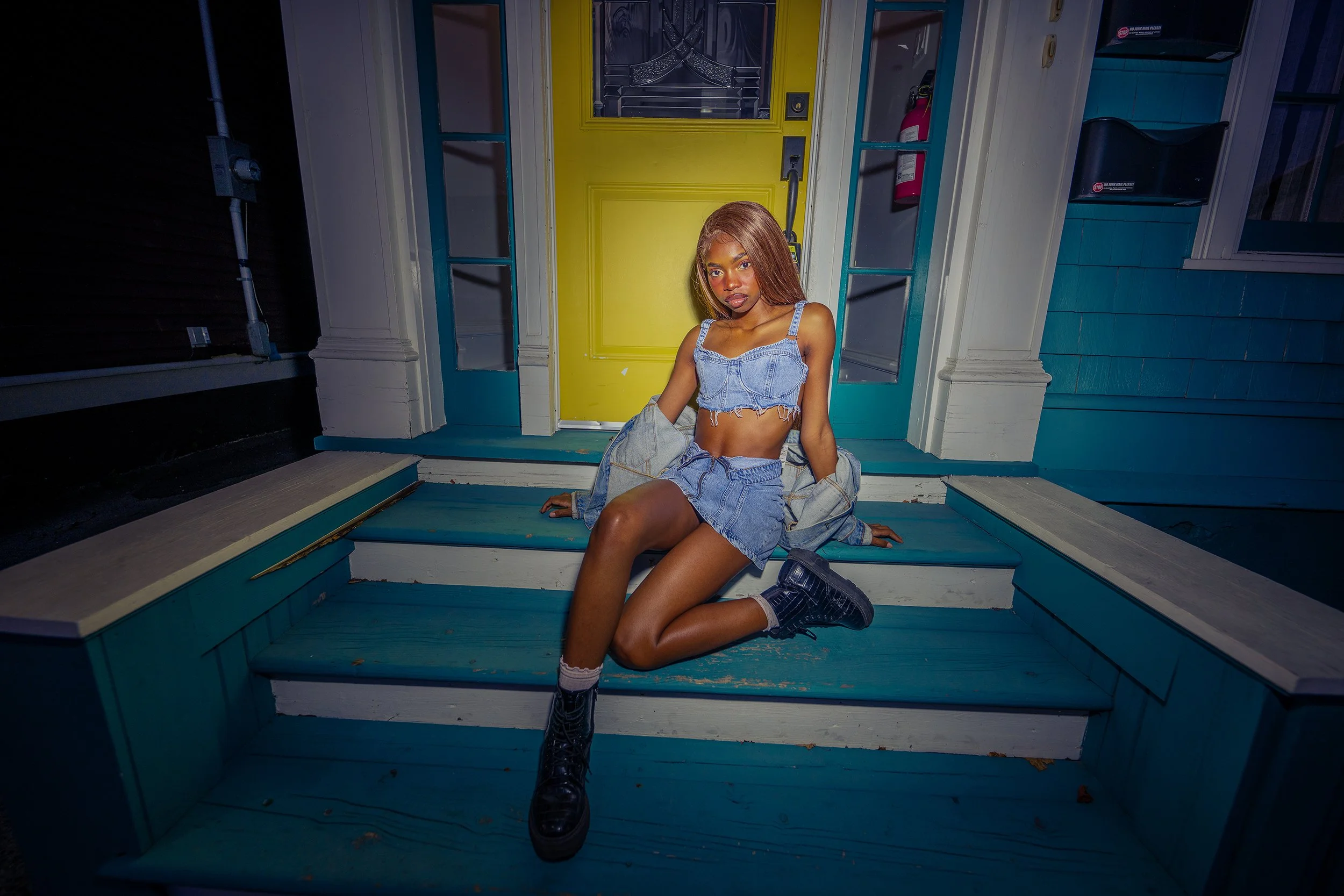 Young woman in denim outfit sitting on porch stairs in front of a yellow door at night
