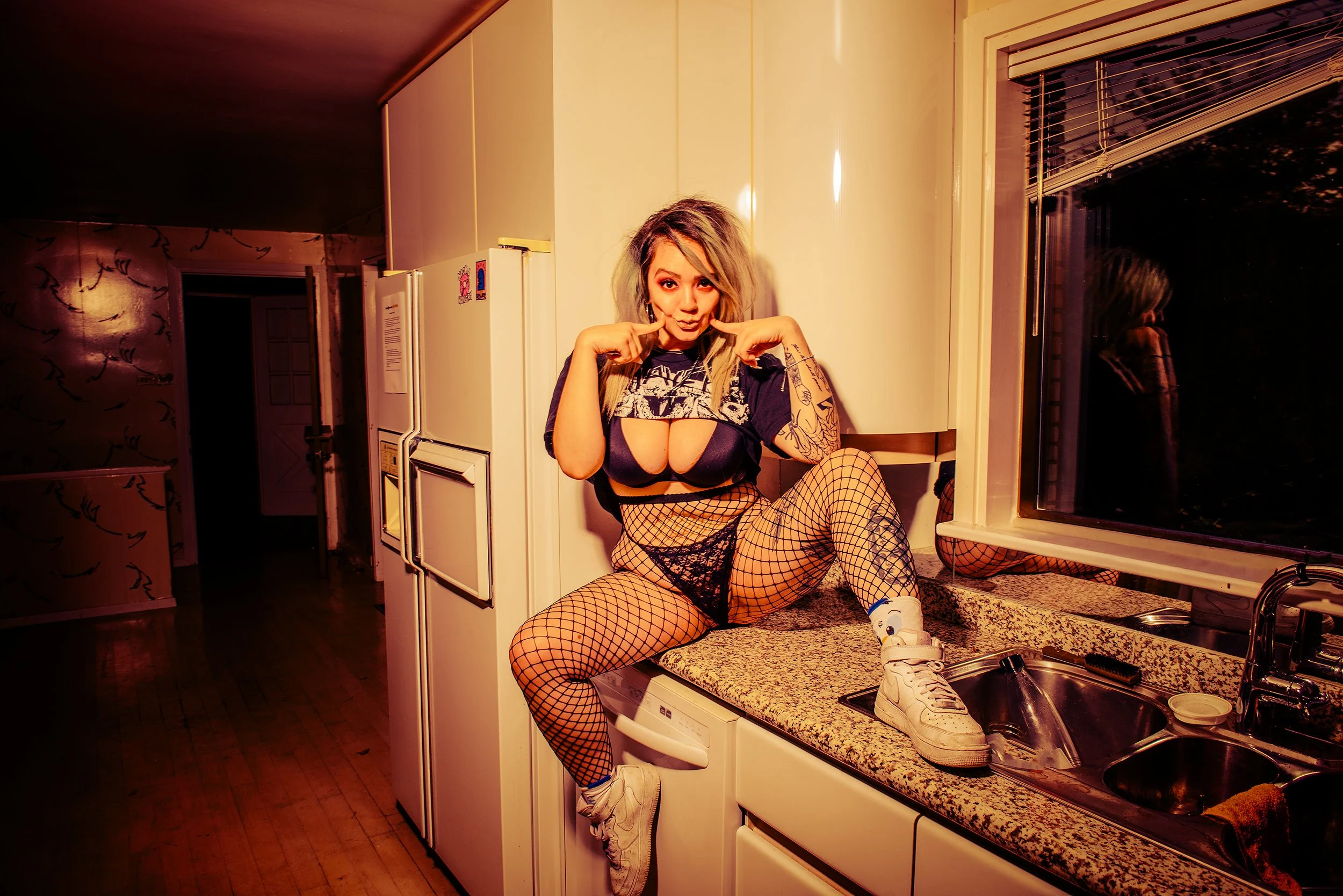 A woman with blonde hair, tattoos, and fishnet stockings sits on a kitchen counter, wearing a crop top, shorts, and sneakers, pointing at her face with her fingers.