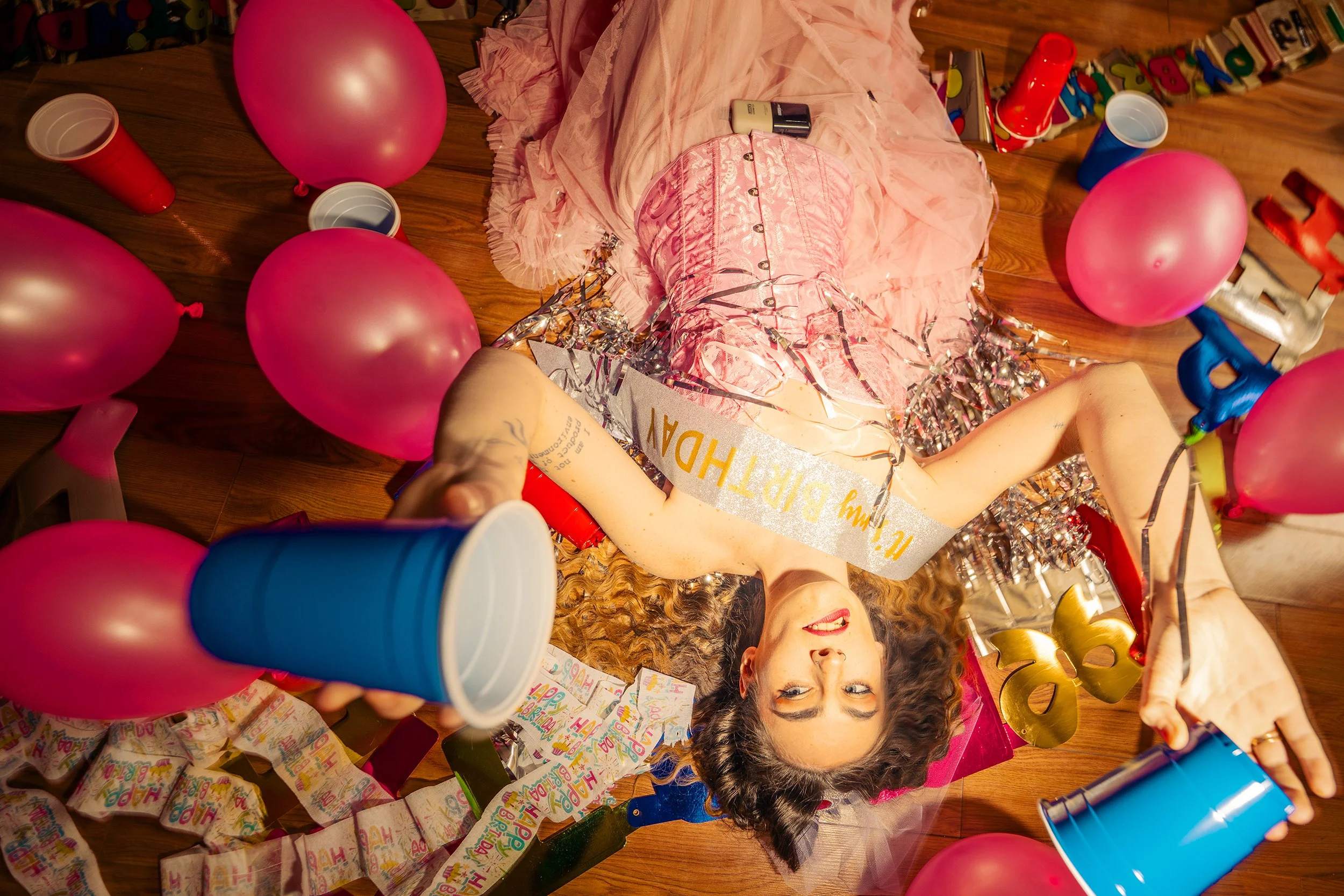 A woman with curly hair is lying on the floor at a birthday party, surrounded by pink balloons, party hats, confetti, and birthday decorations, smiling and wearing a pink dress.