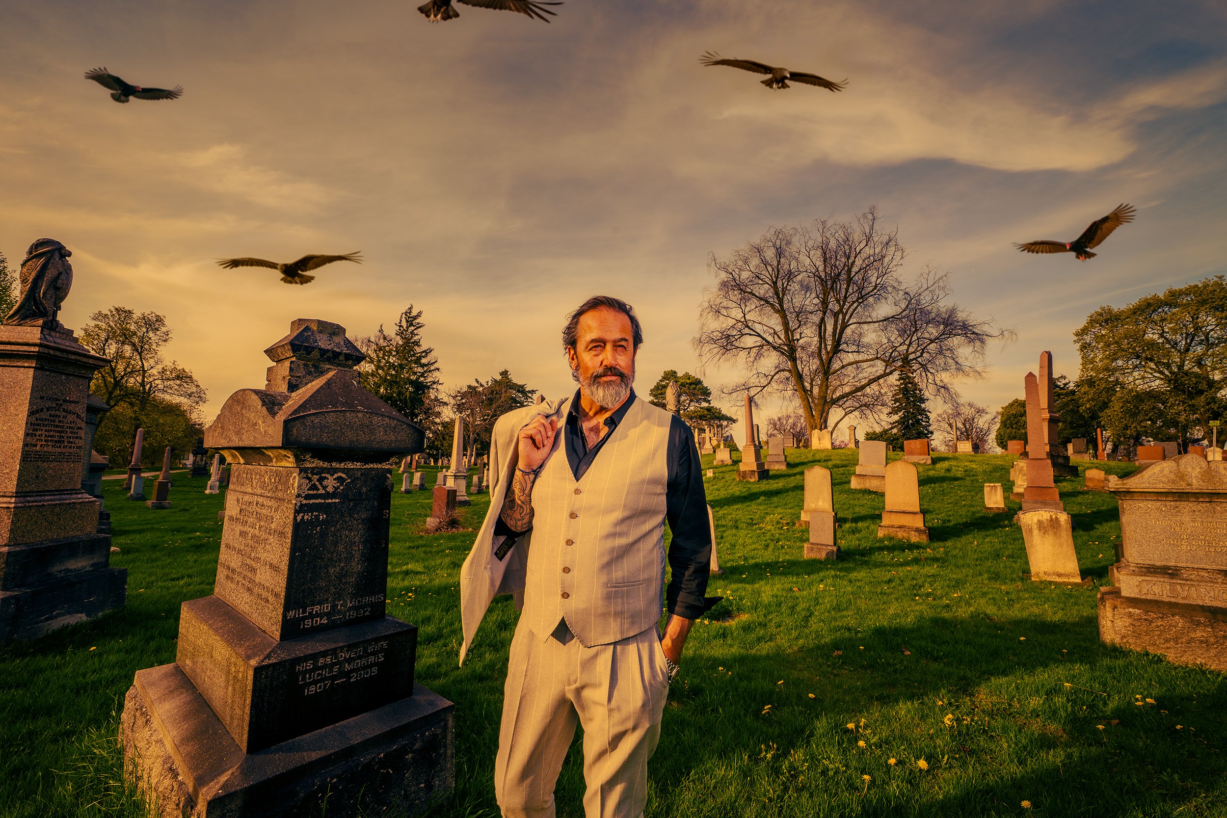 A man with gray hair, beard, and tattoos, wearing a beige vest over a black shirt, standing in a cemetery with tombstones and trees, with birds flying in the sky at sunset.
