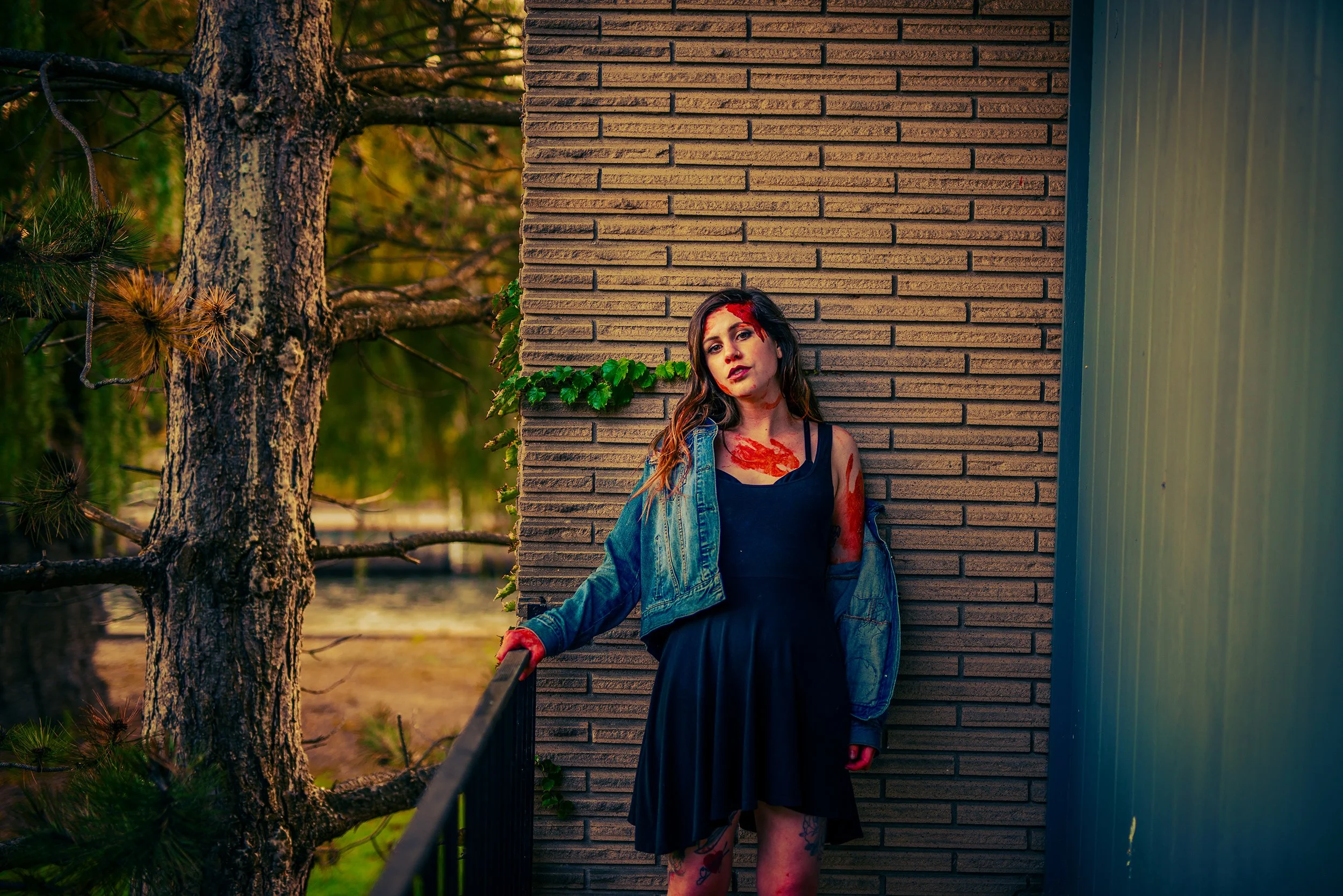 A woman with long hair and tattoos on her legs standing against a brick wall near a green fence, with a large tree in the background. The woman has red paint on her face and body, and is wearing a black dress and denim jacket. The setting appears to 