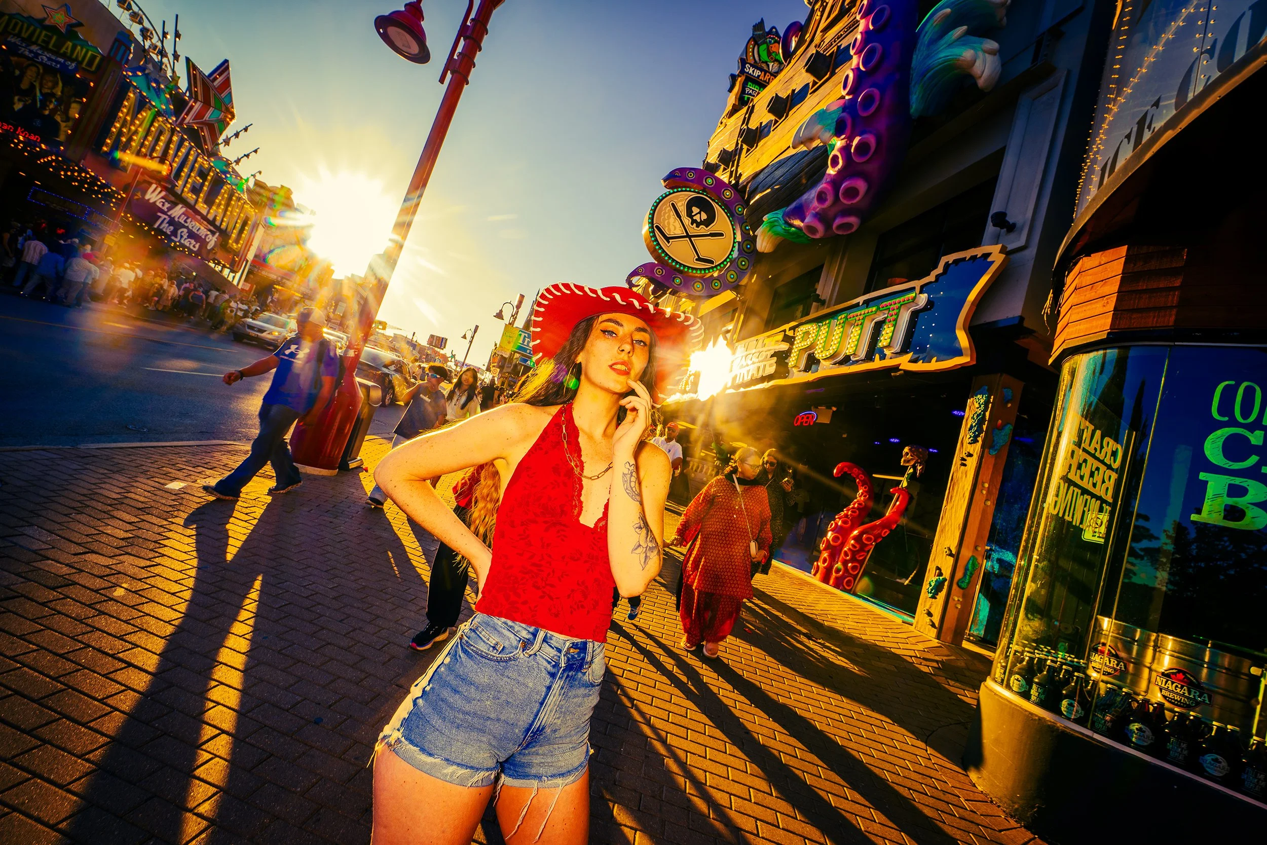 A woman in a red sleeveless top, ripped denim shorts, and a red hat stands on a busy street at sunset, with colorful carnival rides and signs around her.