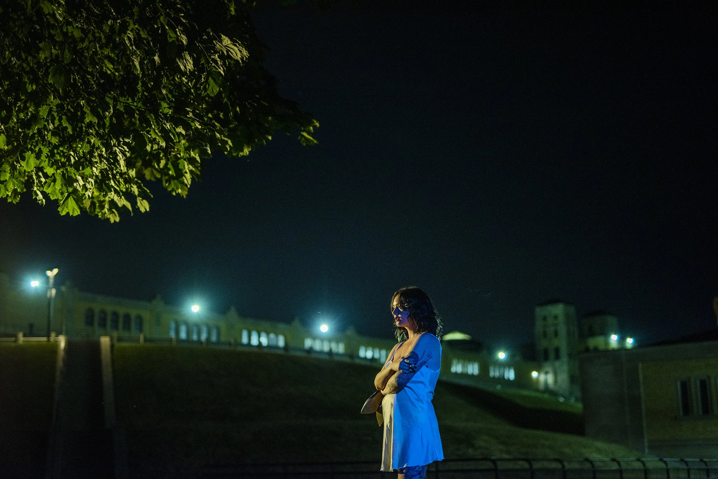 A woman with dark hair and sunglasses stands alone at night on a grassy area near a black metal fence, with a large building and trees illuminated by green and white lights in the background.