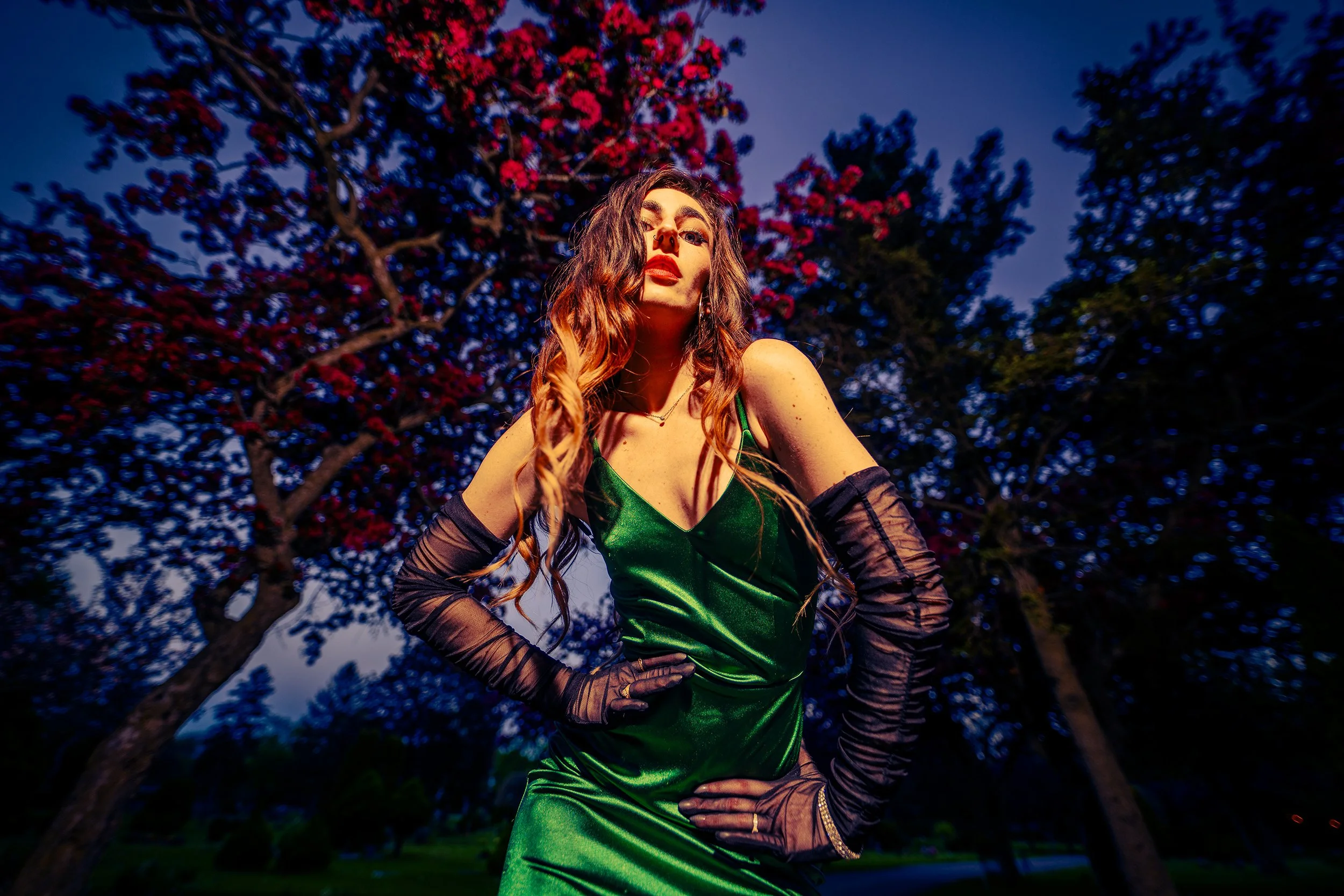 A woman with long wavy hair wearing a shiny green dress and long black gloves stands outdoors in front of a tree with red leaves during dusk.