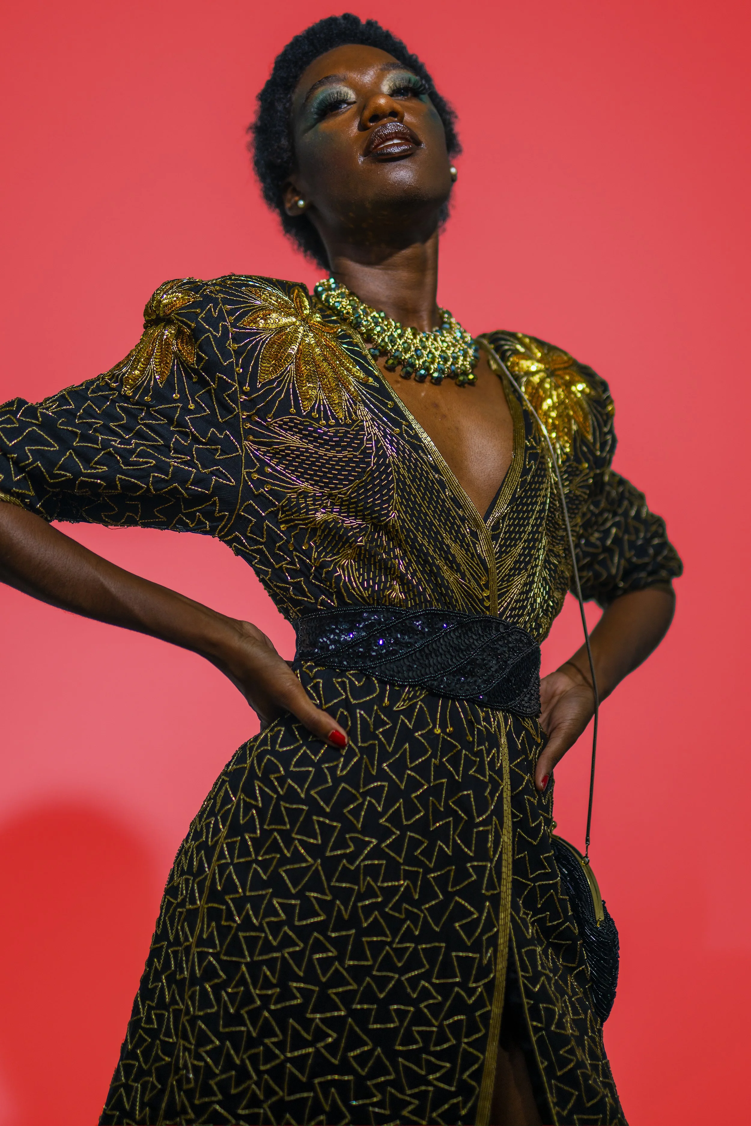 A woman dressed in a black and gold embroidered outfit with a dramatic neckline, wearing a beaded necklace, and standing against a pink background.