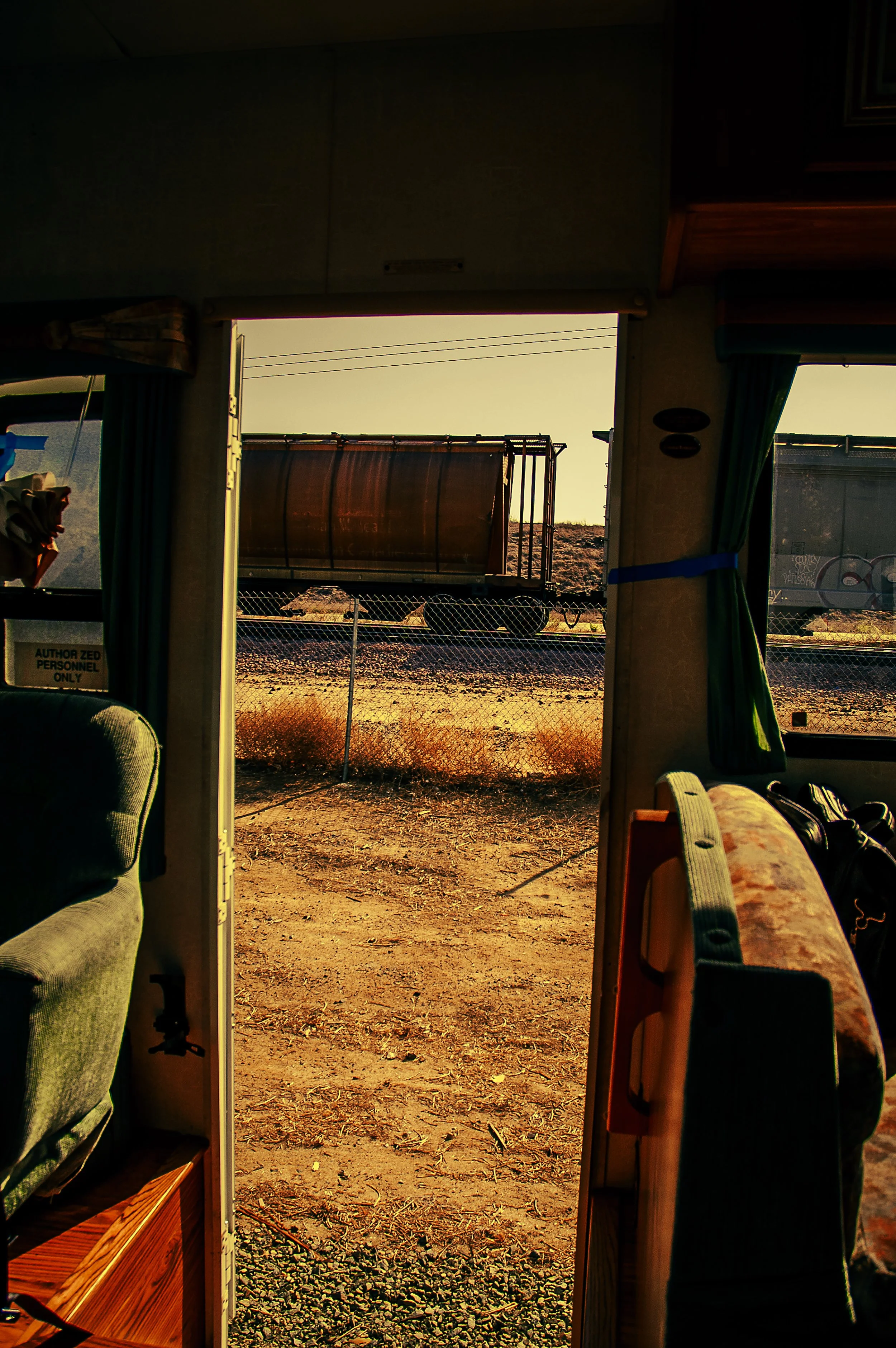 View through a doorway revealing a train car window with a freight train outside at sunset, with dry grass and a chain-link fence in the foreground.