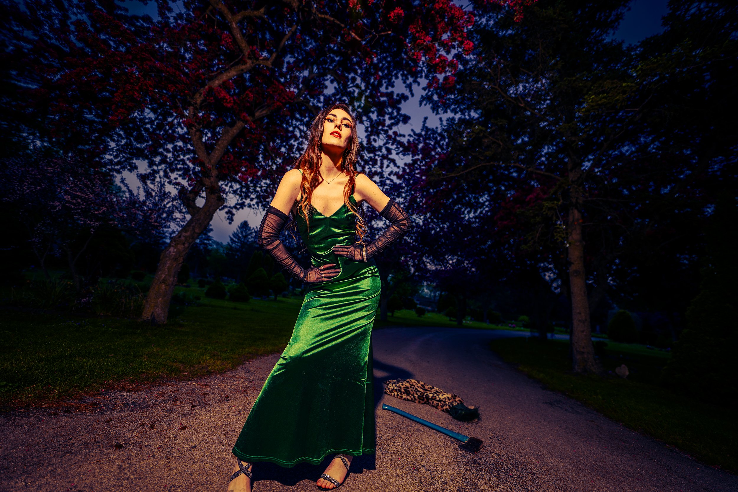 Woman in a green satin dress and black lace gloves standing outdoors at dusk, with trees and a dark sky in the background.