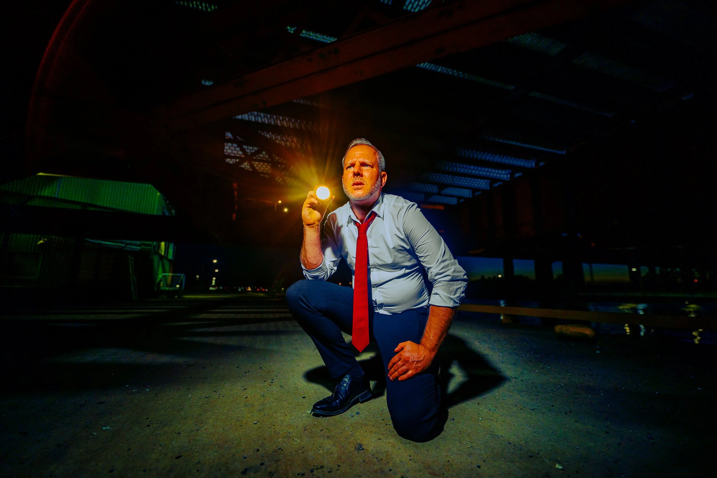 A man wearing a white shirt, red tie, and dark pants is crouching on one knee under a bridge at night. He is holding a flashlight in his right hand, shining light upward, with an intense expression on his face. The background shows the dark underside