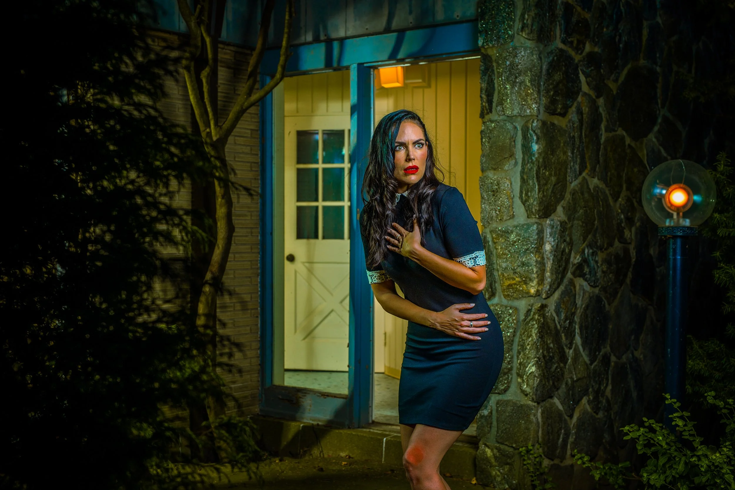 A woman with long dark hair, wearing a fitted black dress with lace sleeves, stands outdoors at night in front of a stone wall and a yellow door. She has a surprised or scared expression, with her right hand on her chest and her left arm bent across 