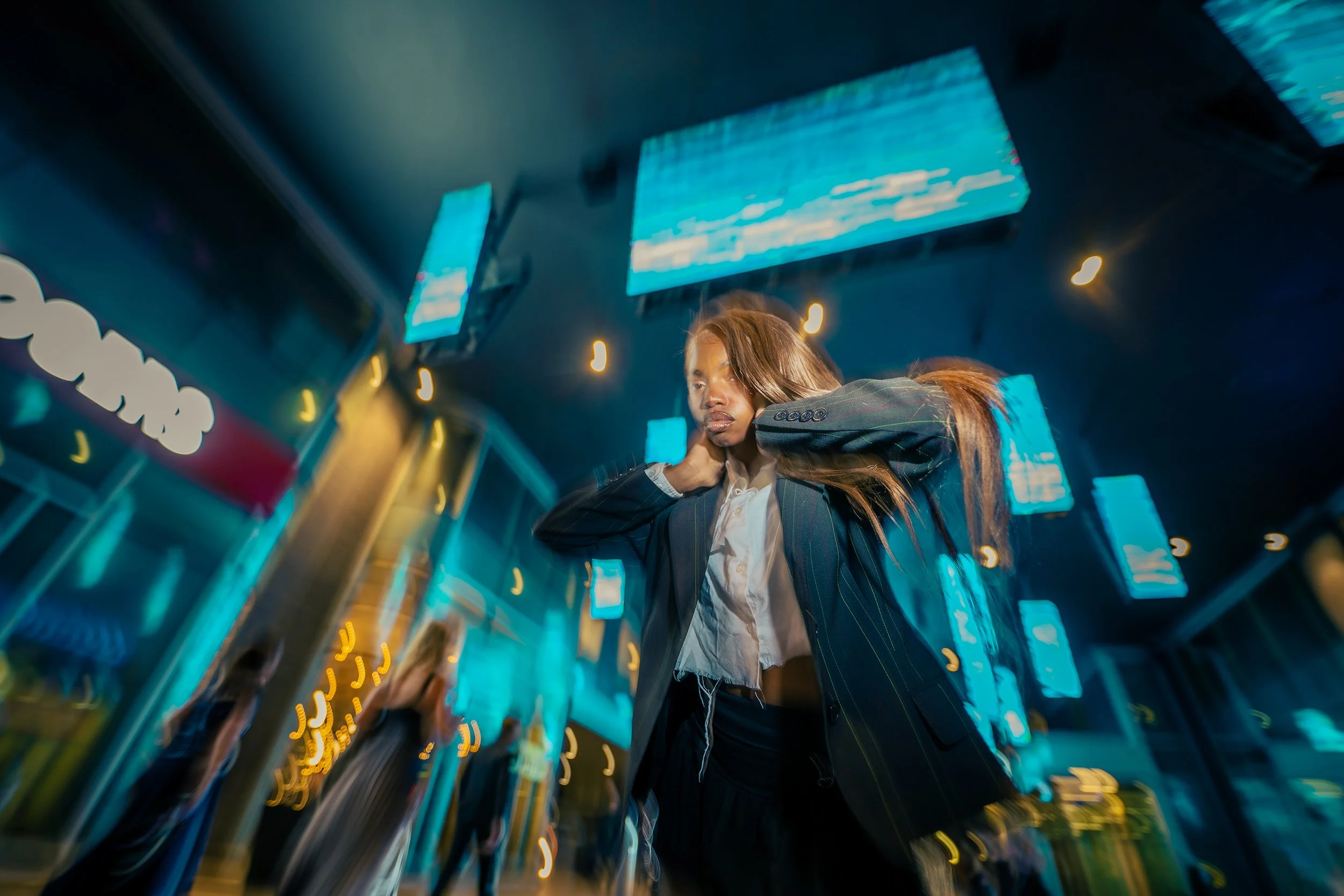 A woman with brown hair, dressed in a pinstripe blazer and white shirt, adjusting her hair in a busy city street at night illuminated by neon lights and digital billboards.