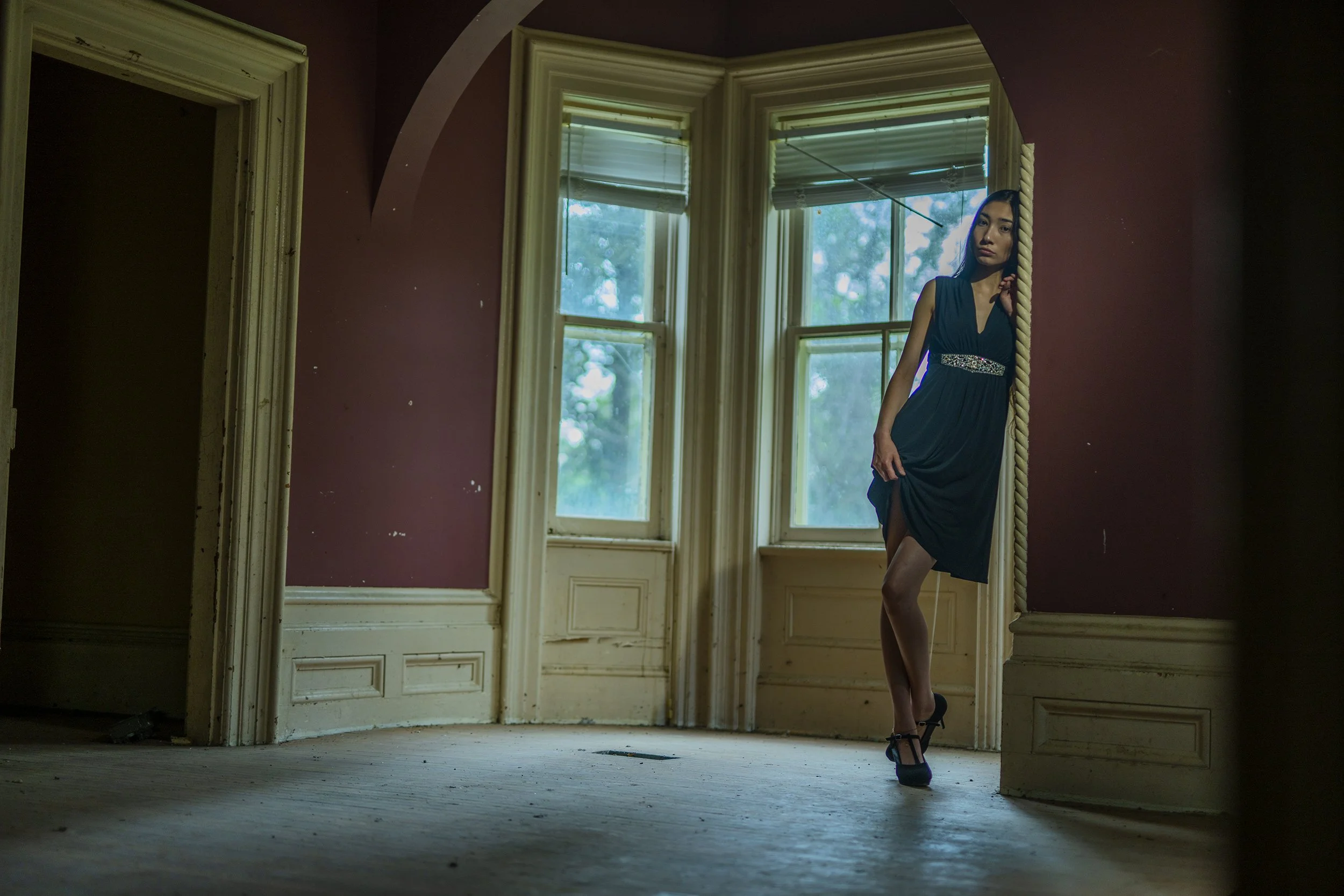 A woman in a dark dress and high heels standing by a large mirror in an old room with tall windows and peeling paint.