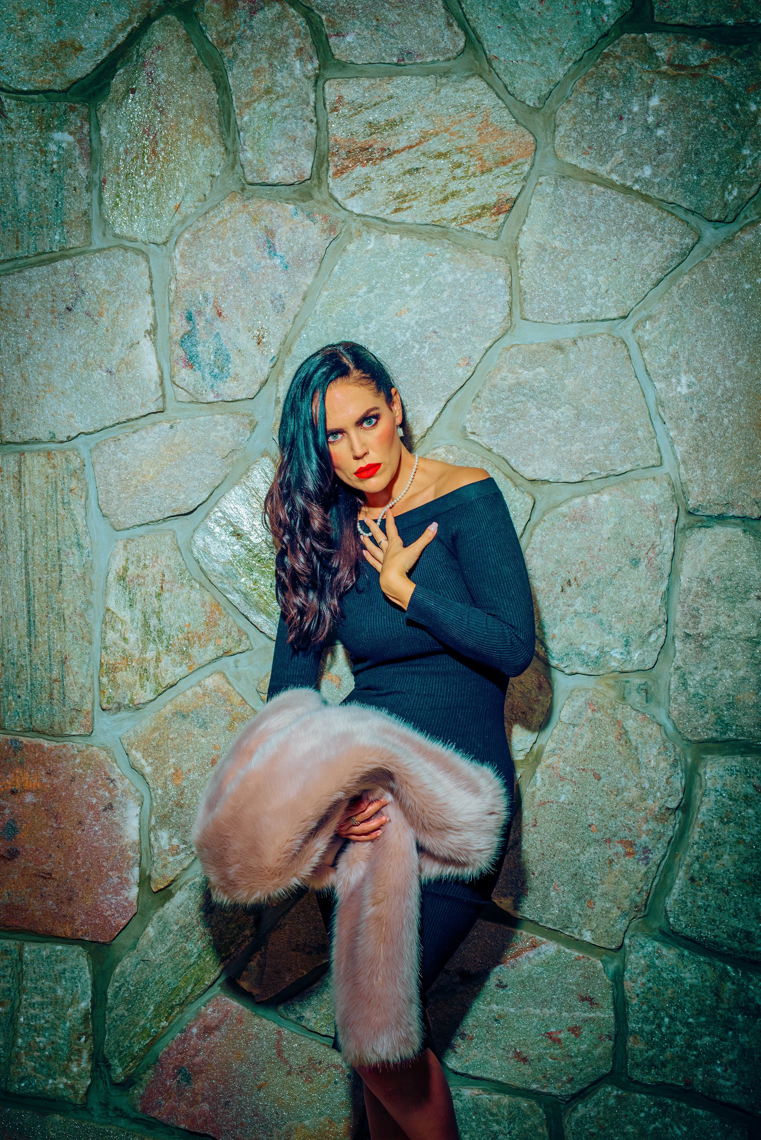 A woman with long dark hair, red lipstick, and earrings, wearing a black off-shoulder top, stands against a stone wall, holding a light-colored fur coat. Part of a person's head is visible at the bottom of the image.