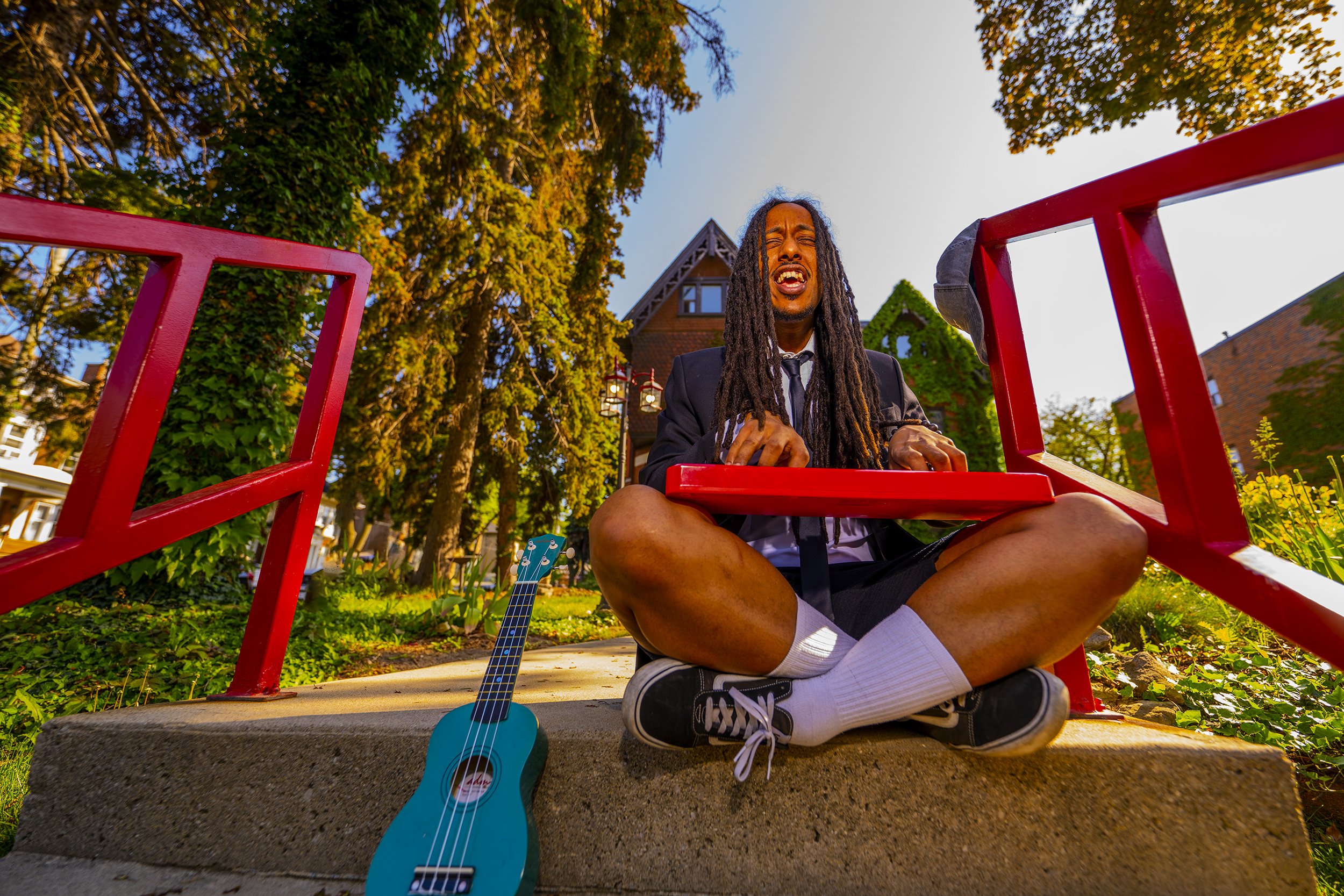 A man with long dreadlocks, wearing a dark blazer and white shirt, sitting cross-legged on the ground with eyes closed and mouth open, outdoors in front of a house with trees, red railing, and a small blue guitar leaning against a concrete block.