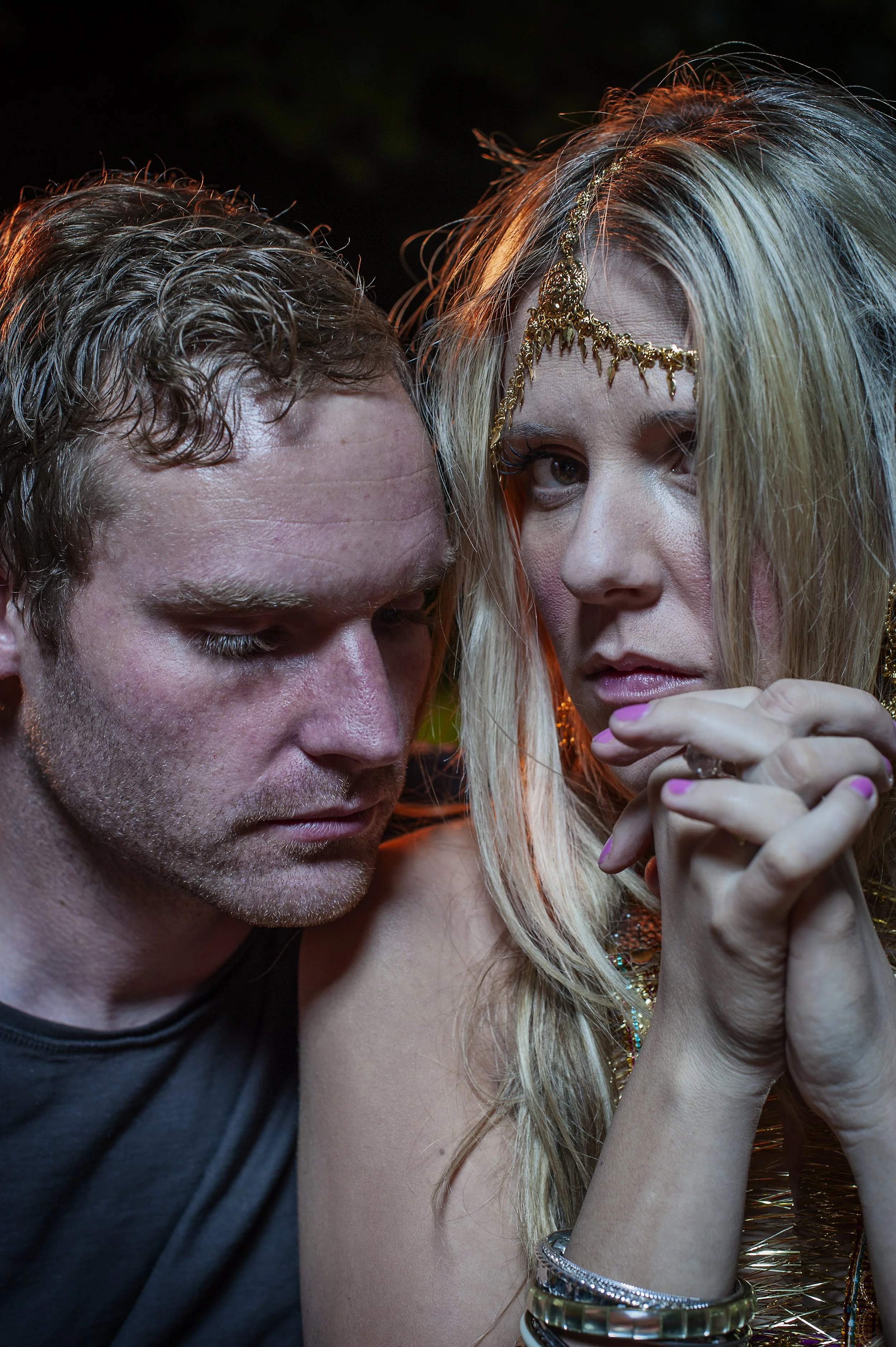 Close-up of a man with wet, curly hair and a woman with long, blonde hair, wearing a gold headpiece and multiple bracelets, with a dark background.