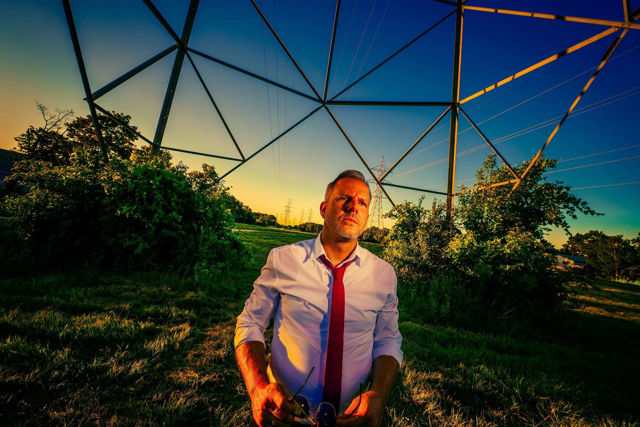 Man wearing a torn shirt and red tie sitting on grass under power lines at sunset.