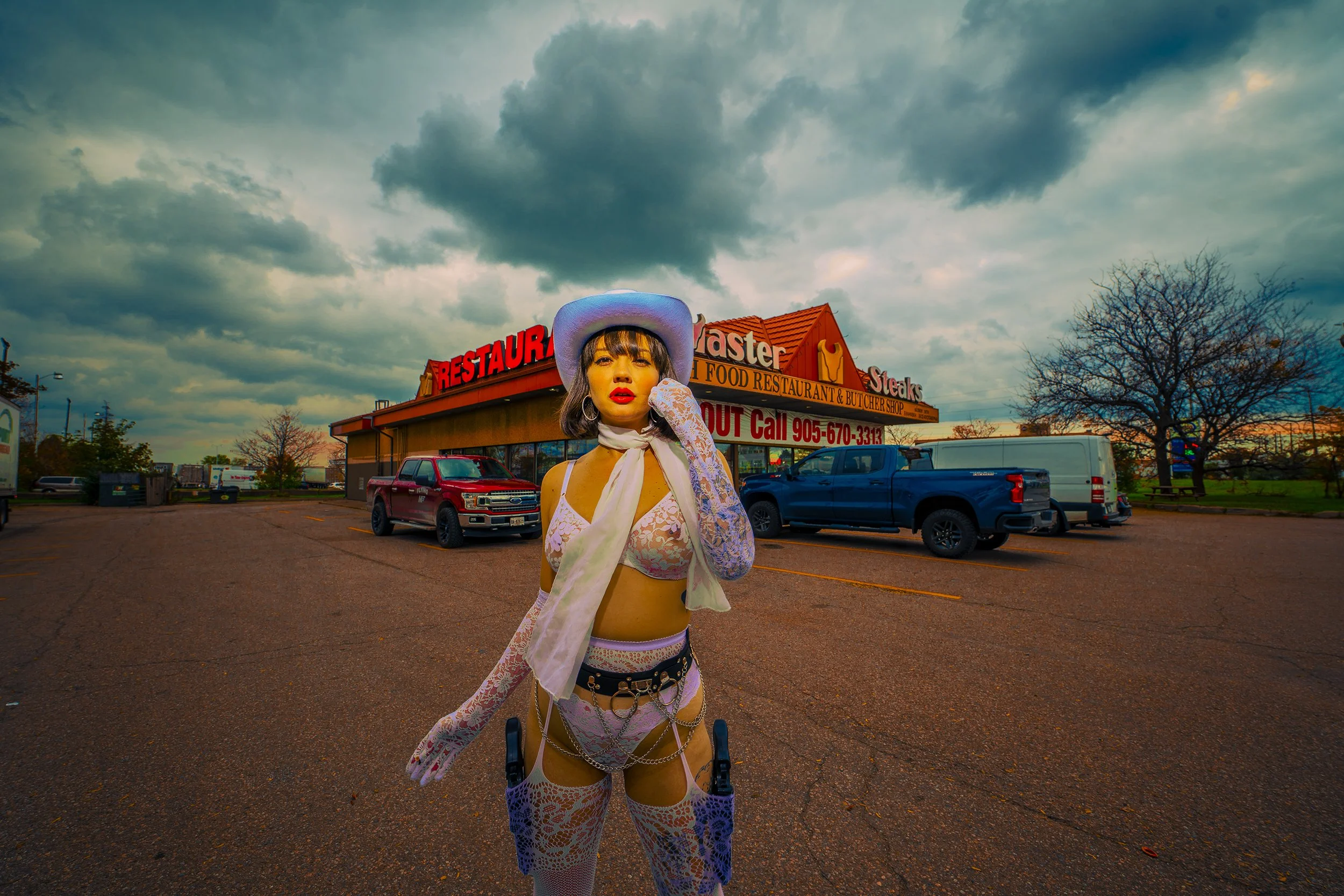 A woman dressed in pink lace lingerie, fishnet stockings, and cowboy hat standing in the parking lot of a restaurant under a cloudy sky.